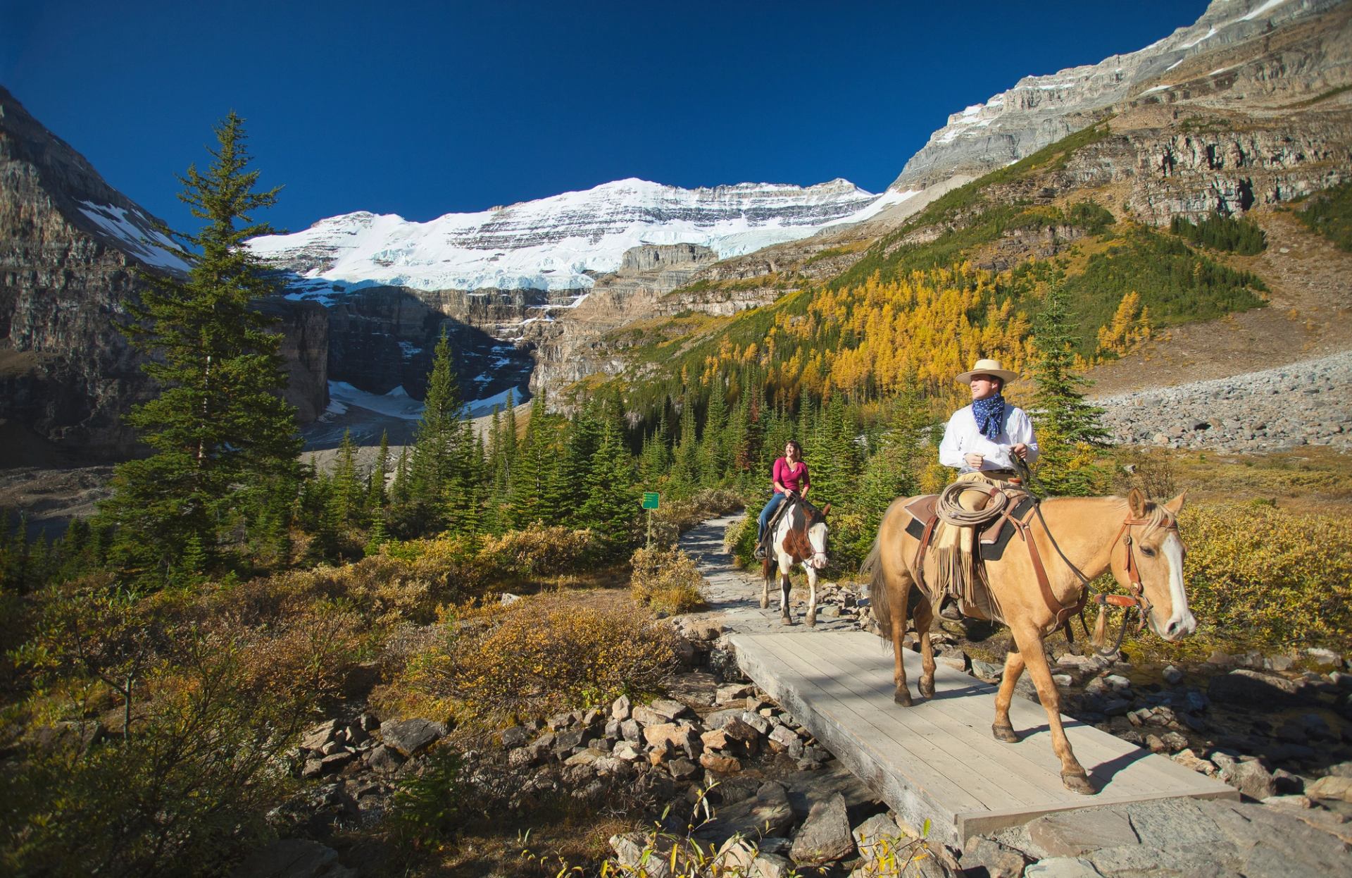 Horseback riders cross a wooden trail bridge below snow-capped mountain and forest.