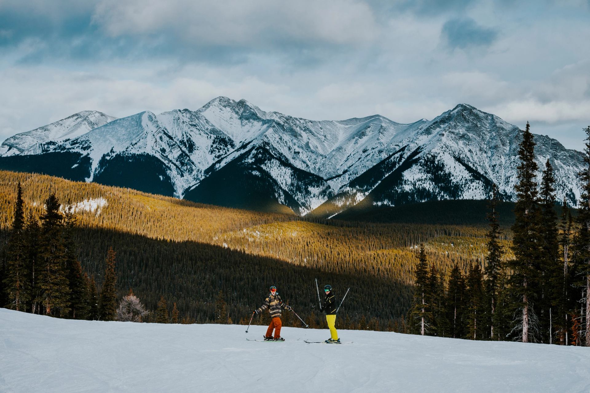 Two skiers with views of the mountains, the sun illuminating the valley, while skiing at Nakiska Ski Resort in Kananaskis Country