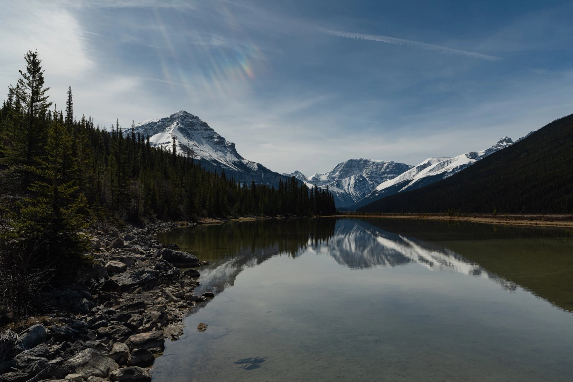 Calm lake reflecting snow-capped mountains and trees under a clear sky with faint rainbow.