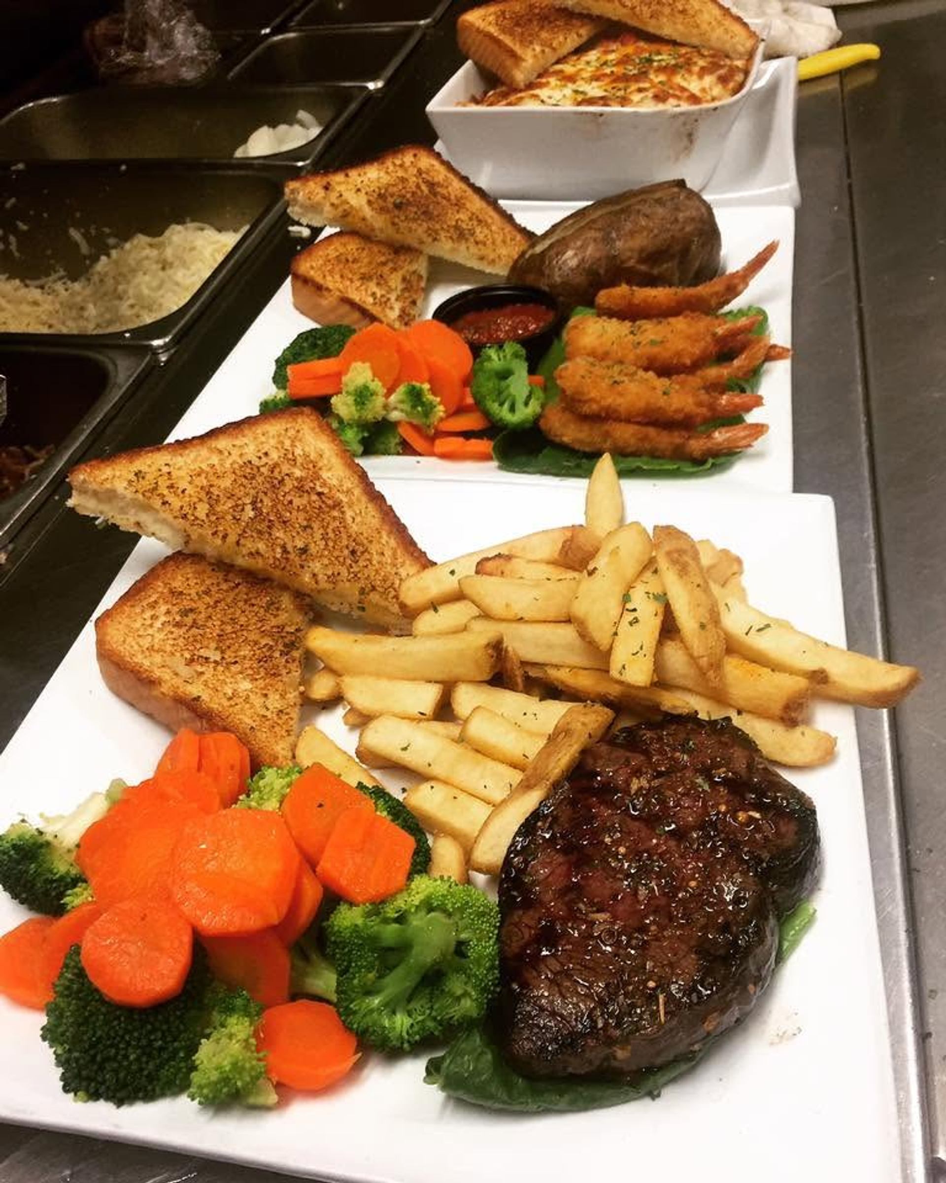 Steak, fries, vegetables, shrimp, baked potatoes, and toast are plated and ready to be served.