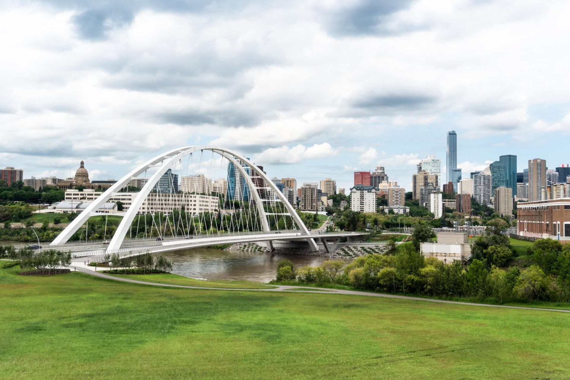 A modern white arch bridge crosses a river, with a city skyline visible behind a large green park under a cloudy sky.