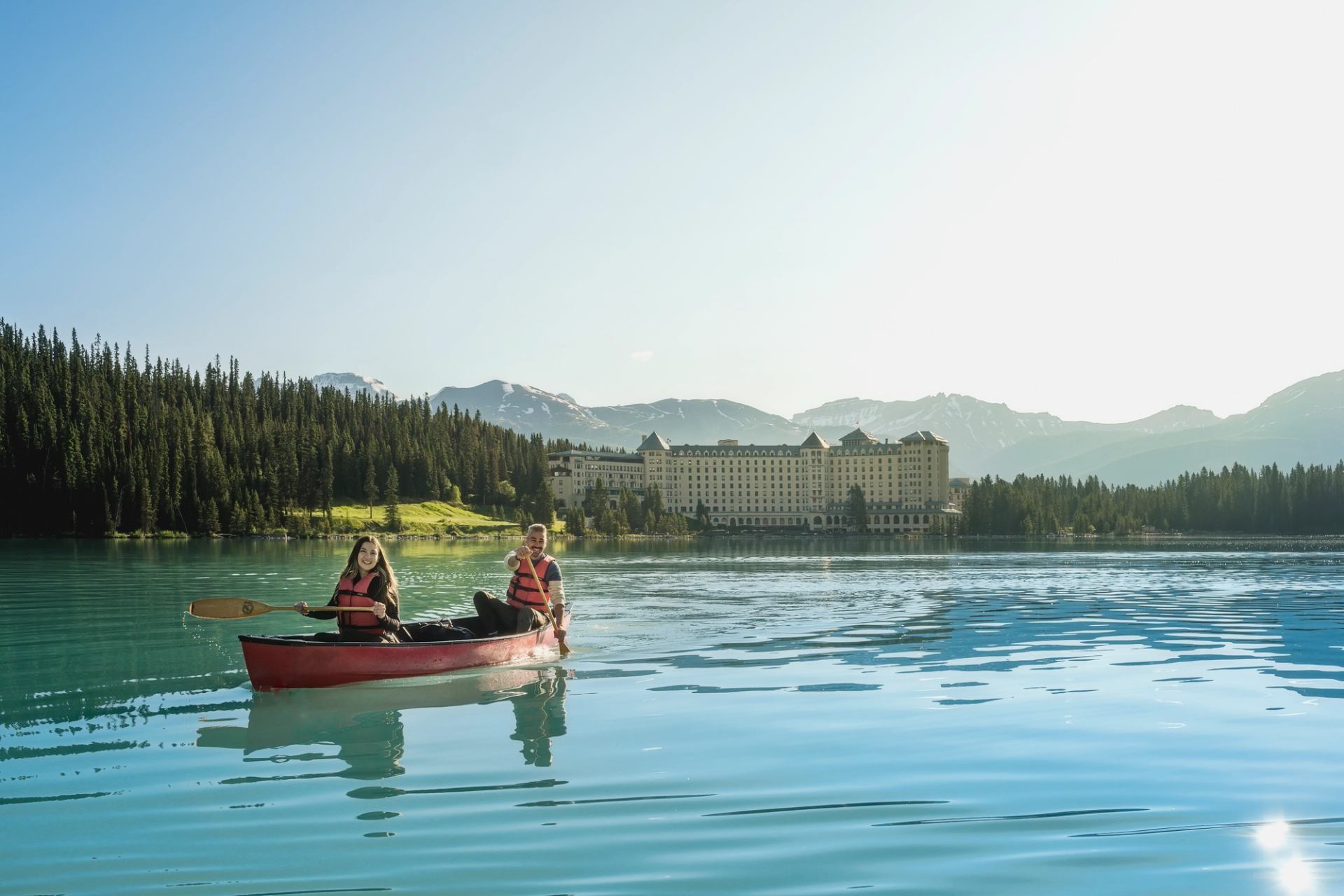 Red canoe on turquoise lake with life-jacketed paddlers, forest, hotel and mountains behind.