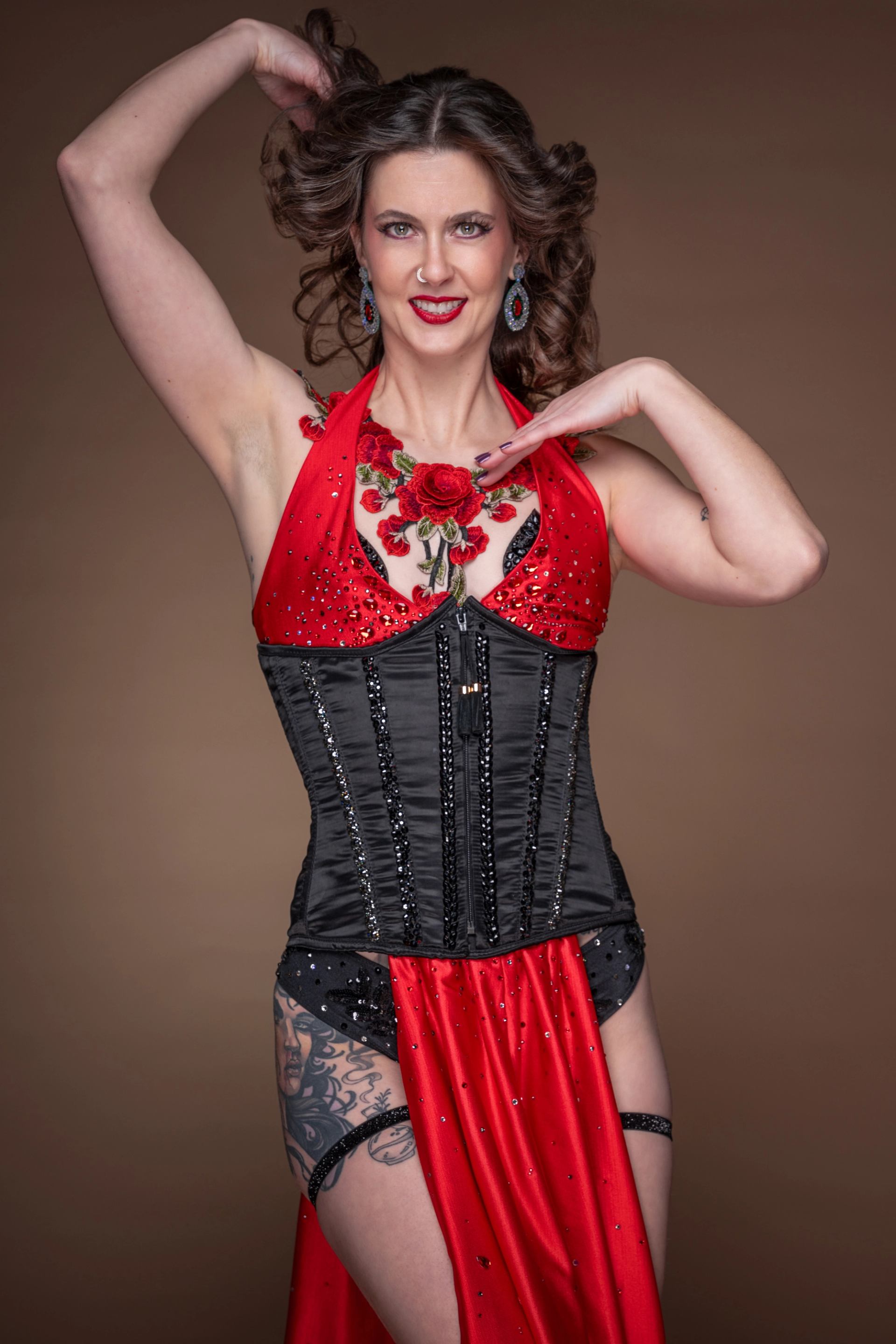 A smiling woman in a sparkling red halter top with rose embroidery, black corset, and a red skirt, posing with her hands raised.