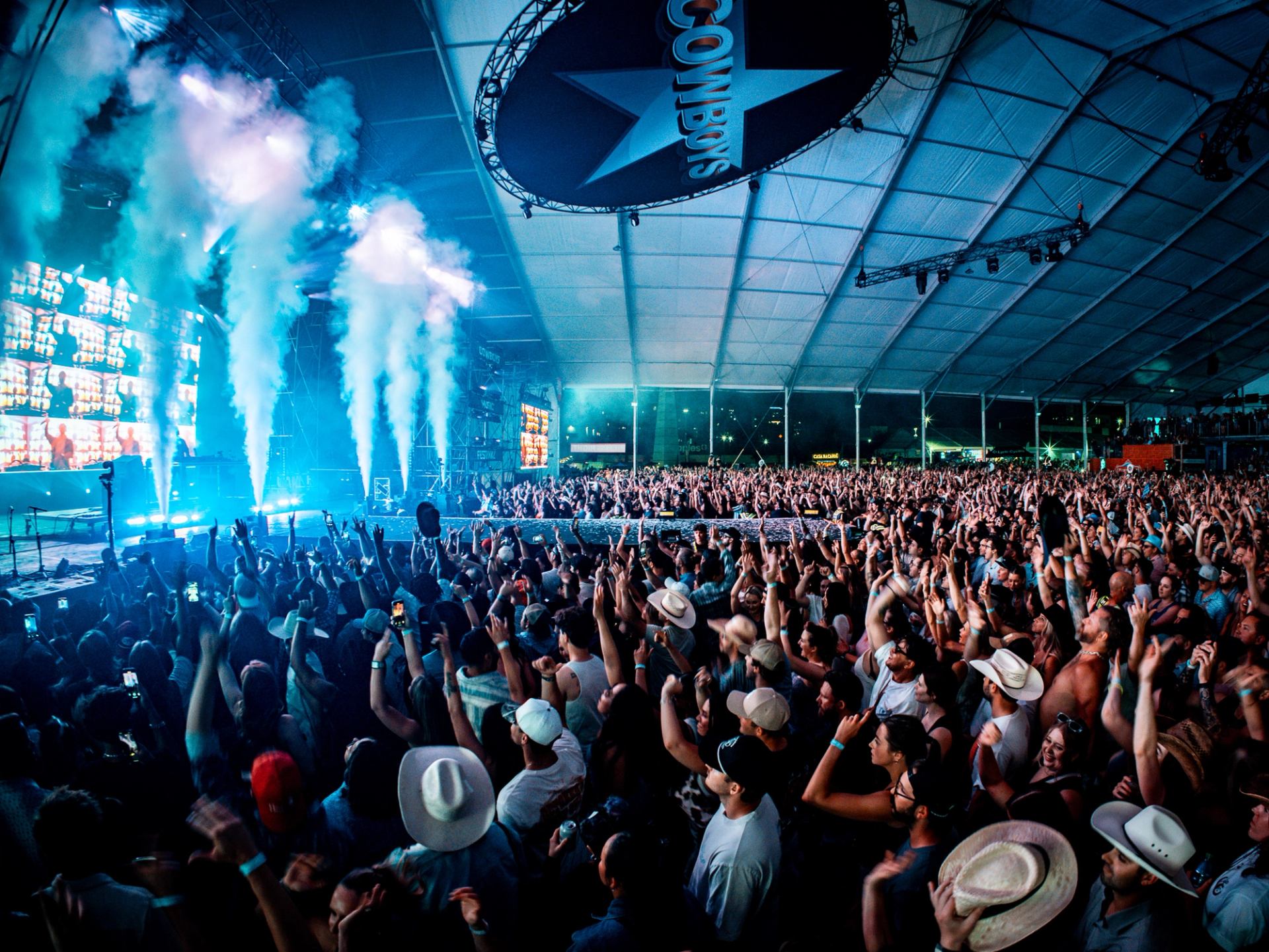 Crowd with raised hands facing stage lights and smoke effects inside the Cowboys Music Festival tent.
