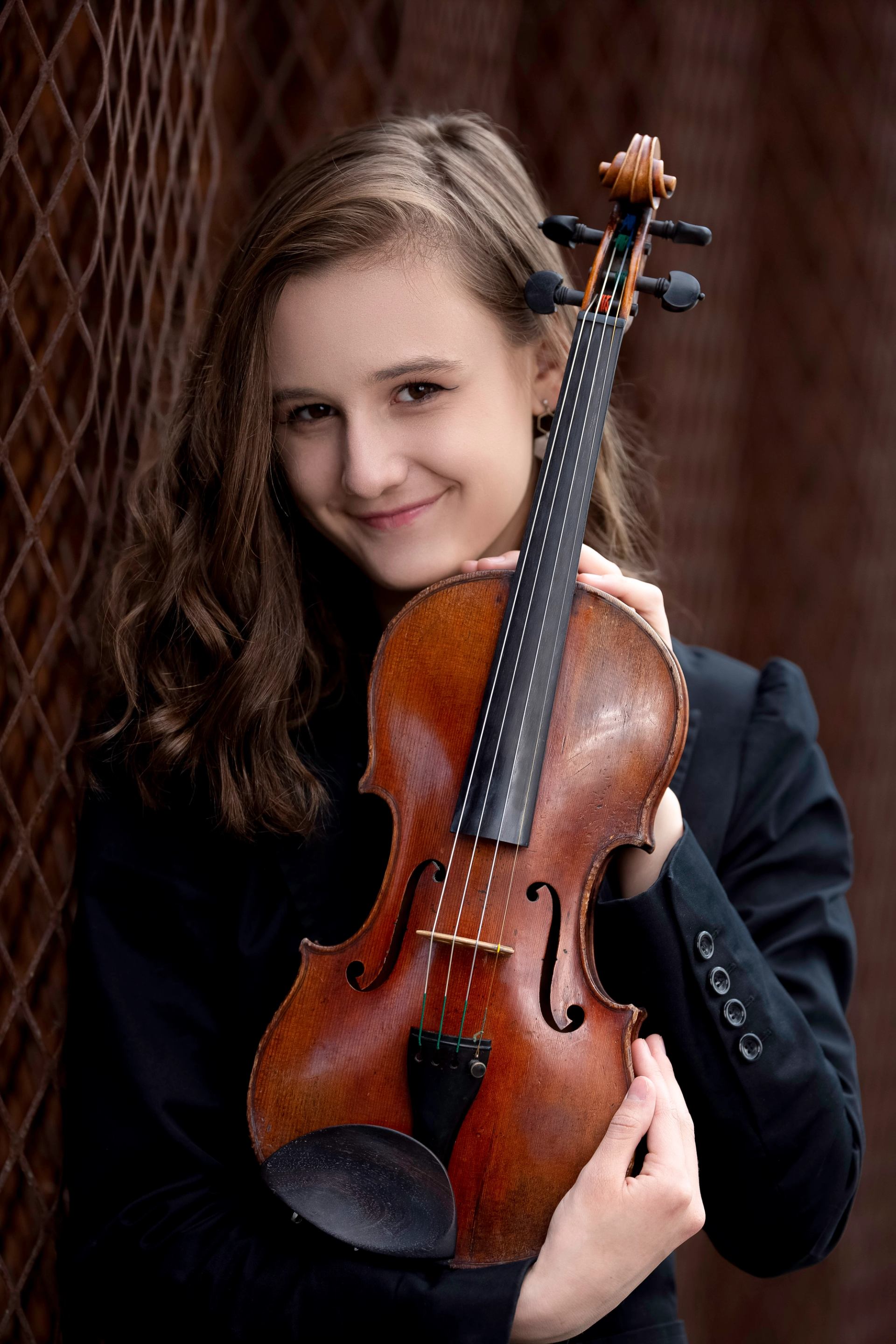 Person in a black jacket holds a wooden violin against a brown metal backdrop.