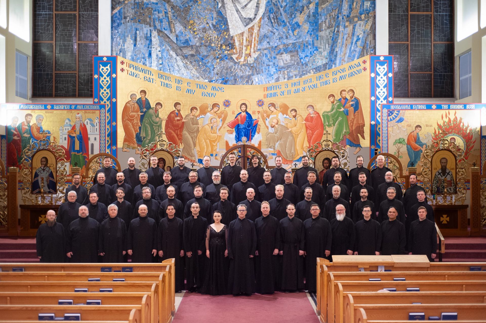 Large choir in black robes stands before colourful Orthodox church iconostasis.