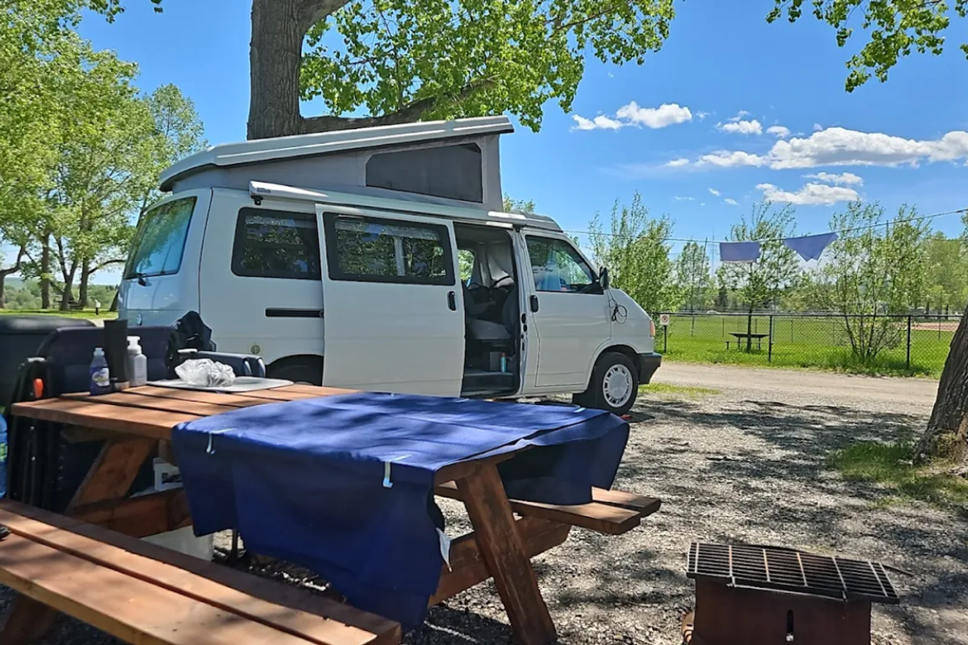 Diamond Valley Municipal Campground features a daytime campsite with a white camper van parked beside a wooden picnic table covered with a blue cloth under leafy trees. The site includes a fire pit and overlooks an open grassy area with fencing and clear blue skies.