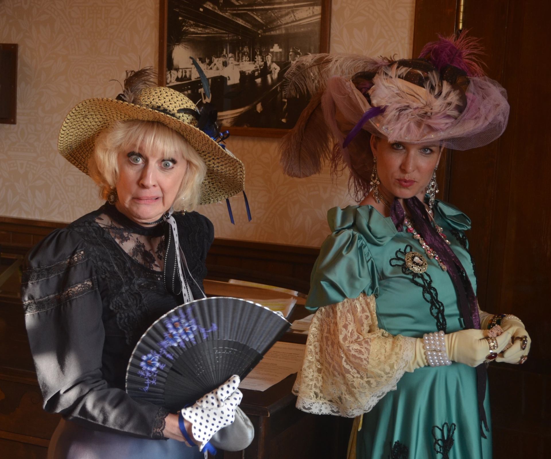 Two people in ornate dresses and feathered hats pose indoors with a folding fan.