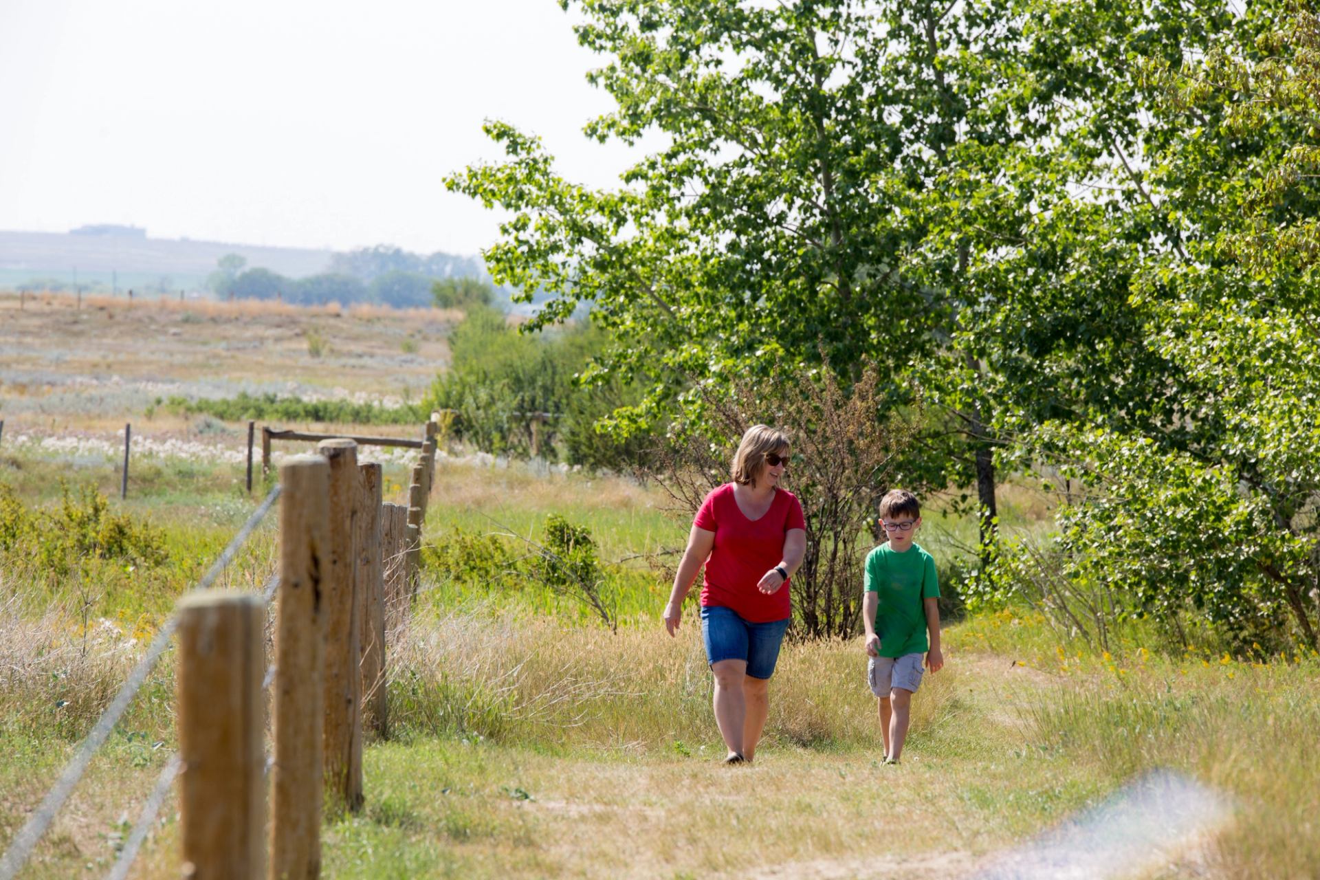 Two people walk along a grassy trail beside a wooden fence and trees.