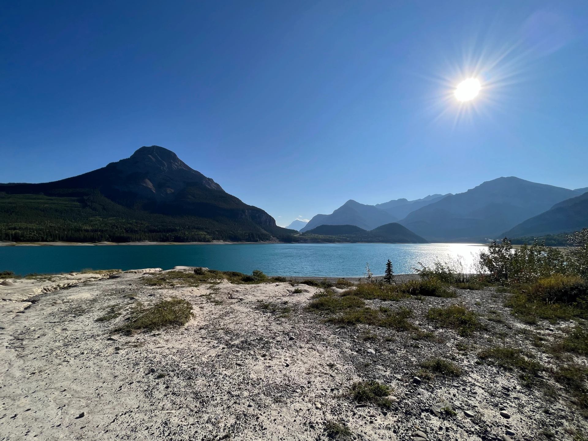 Bright sunlight over a blue mountain lake with rugged peaks behind it.