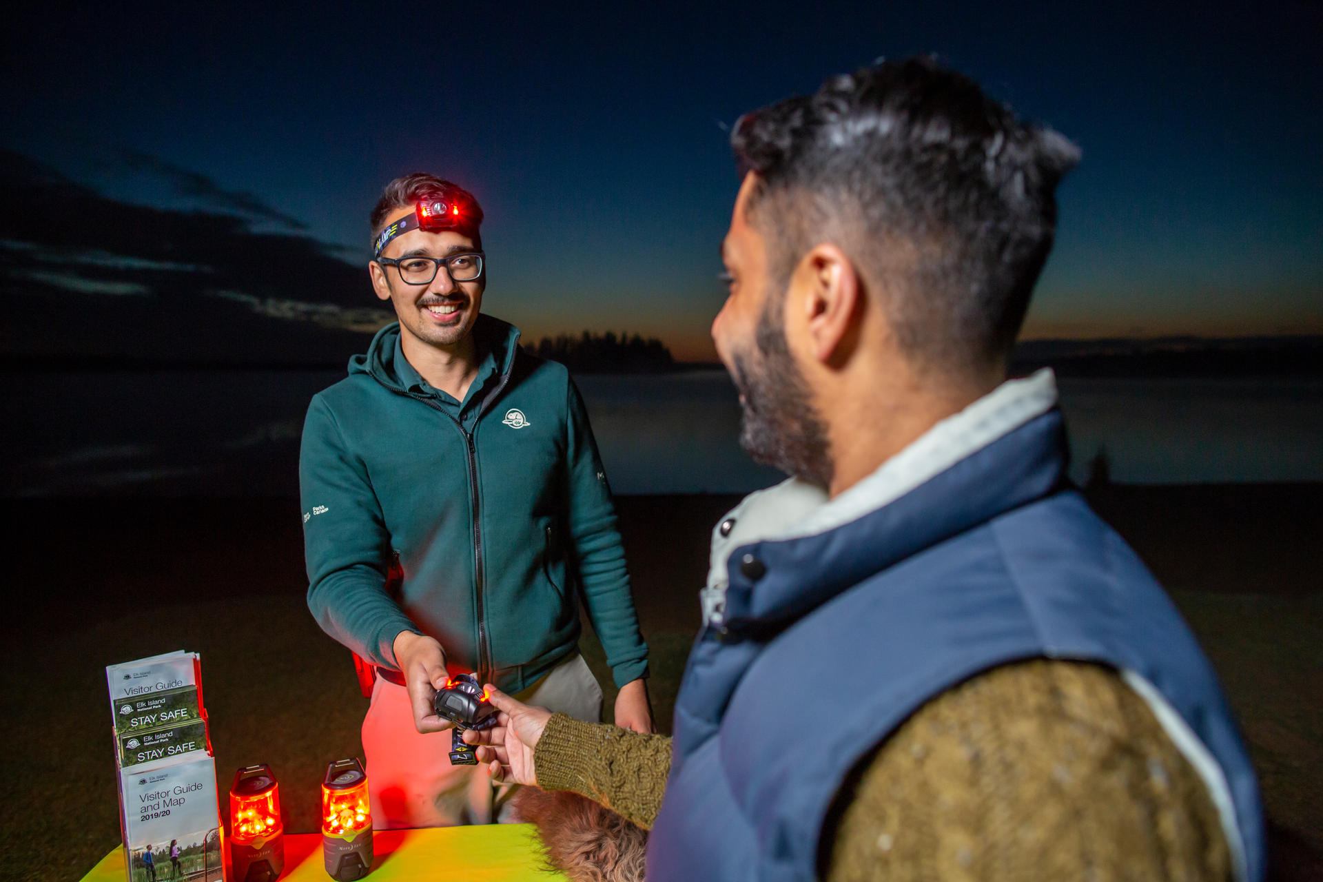 Person wearing a headlamp hands another person a light beside illuminated lanterns.