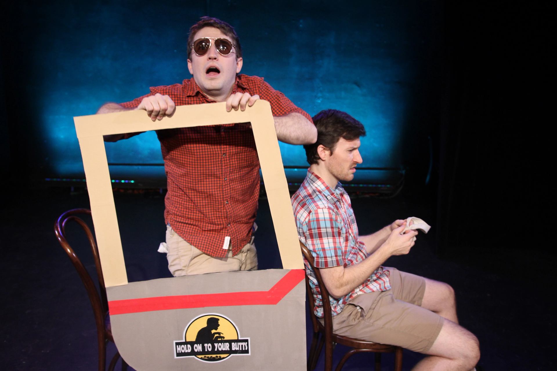 Stage performance with two people miming a car ride using a cardboard frame prop.