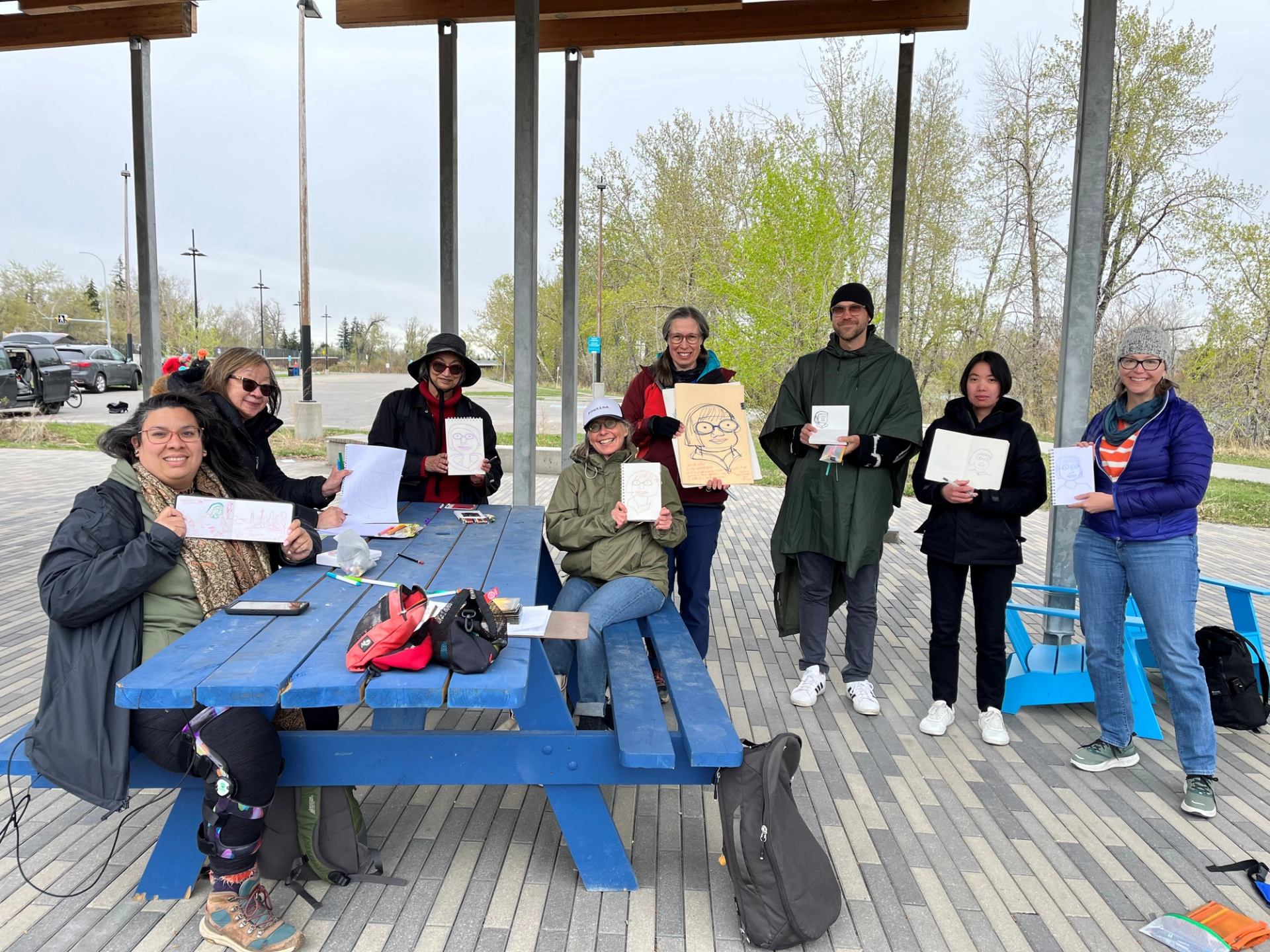 Group of people at blue picnic table holding sketchbooks and drawings outdoors.