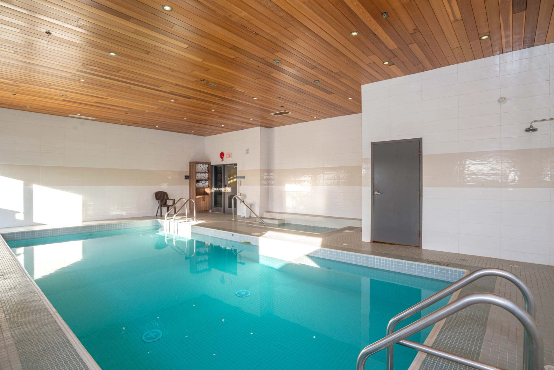 Indoor swimming pool with metal handrails, hot tub, towel shelf and wood-panelled ceiling.