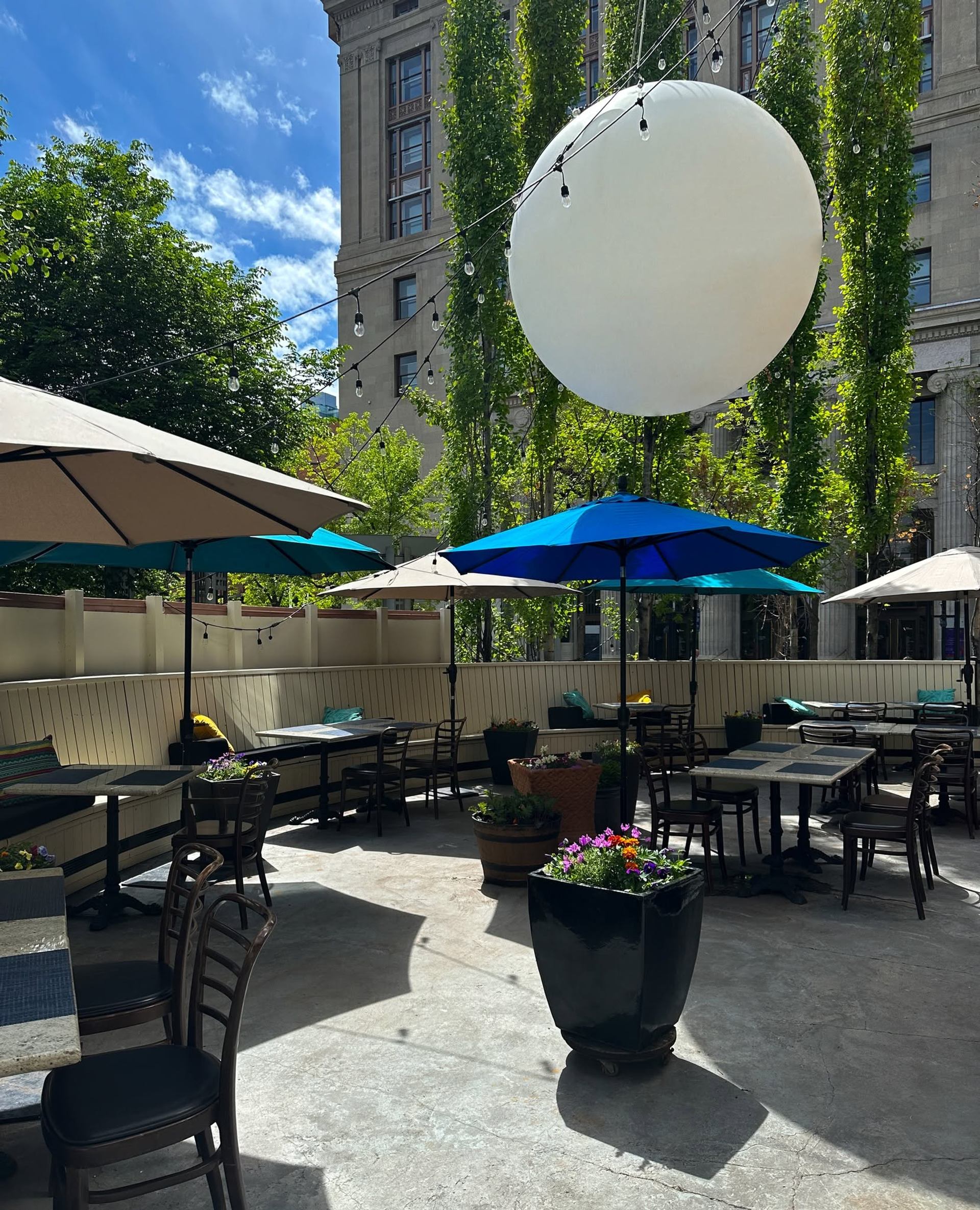 Outdoor patio with tables, umbrellas, string lights and planters beside tall office building.