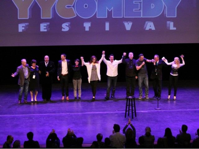 Performers pose with arms raised on stage at a theatre festival, with audience seated.