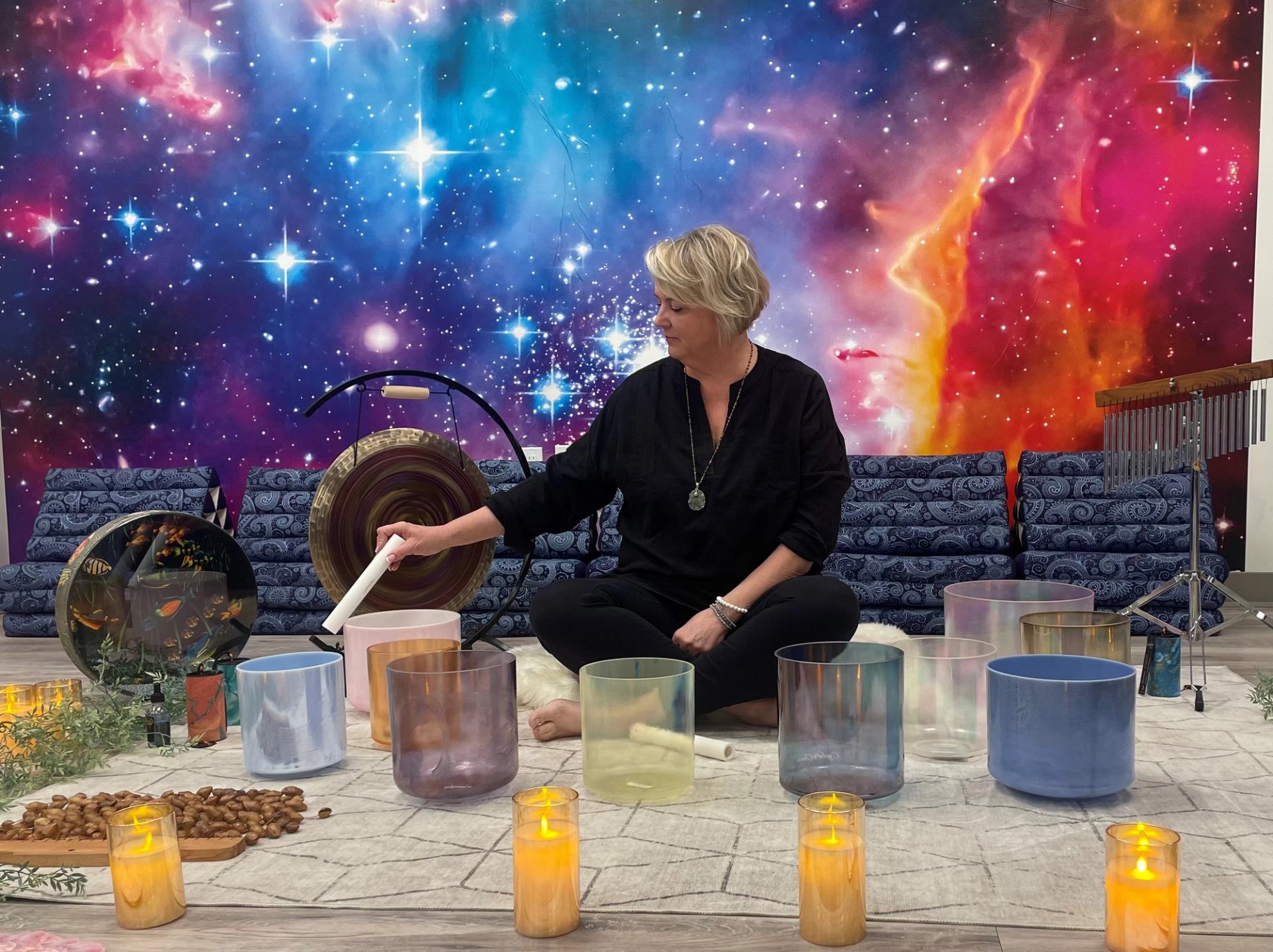 Person seated among crystal singing bowls and candles before colourful space mural backdrop.