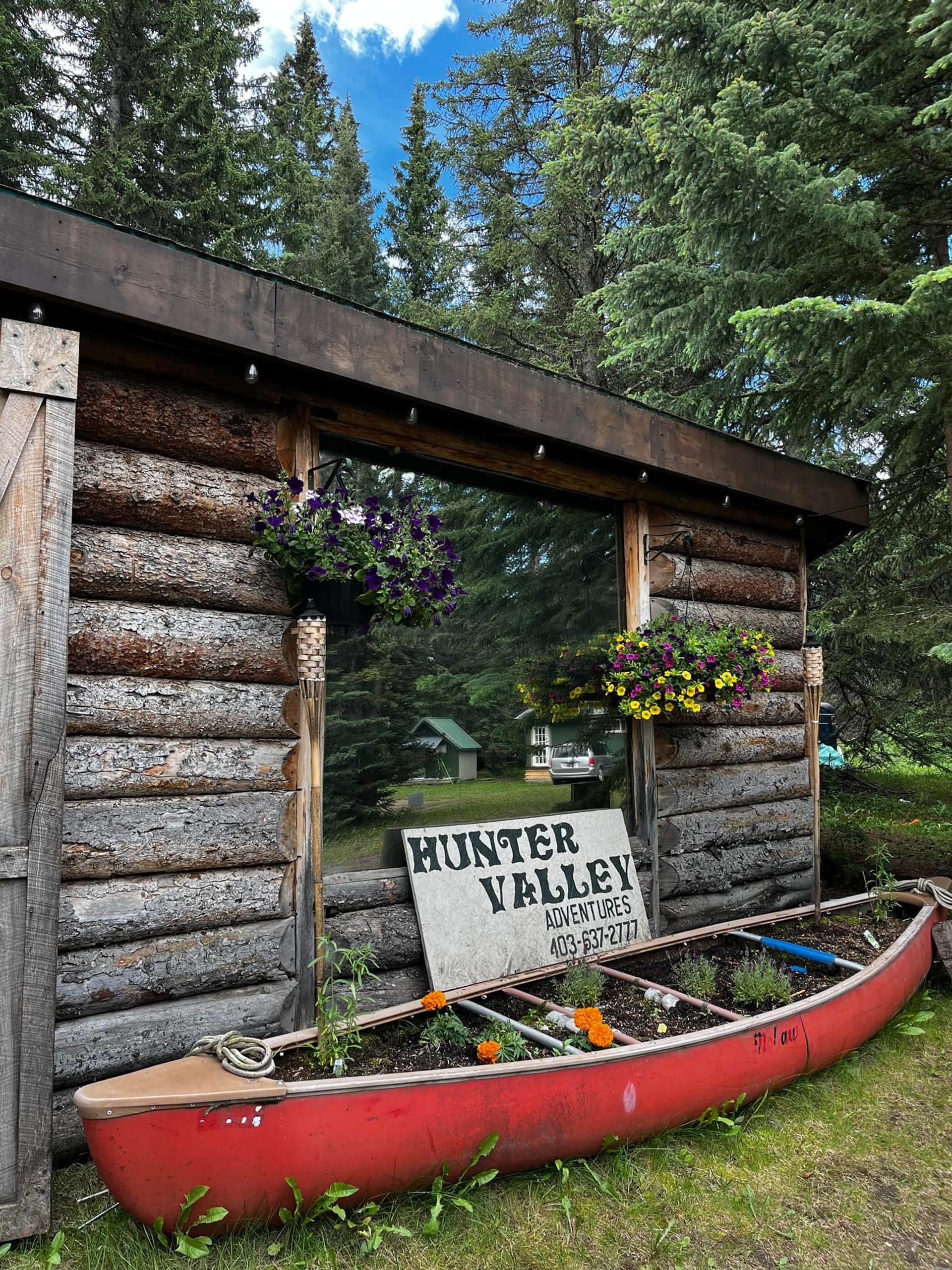 Red canoe planter and Hunter Valley Adventures sign beside rustic log cabin wall.