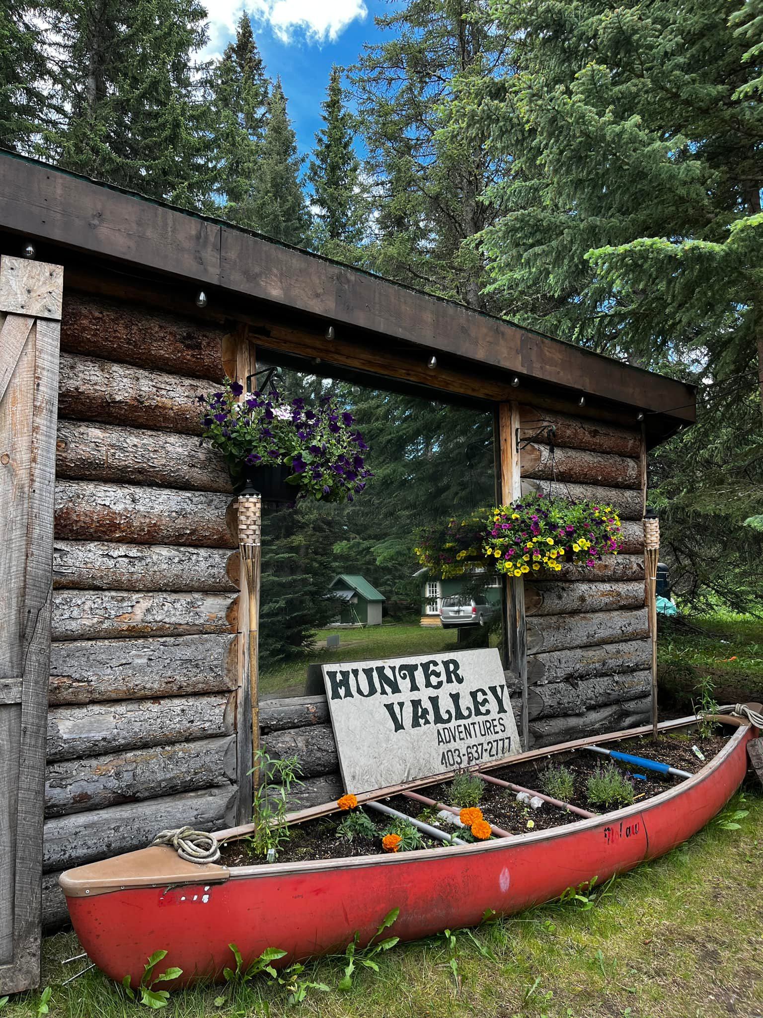 Red canoe planter and Hunter Valley Adventures sign beside rustic log cabin wall.
