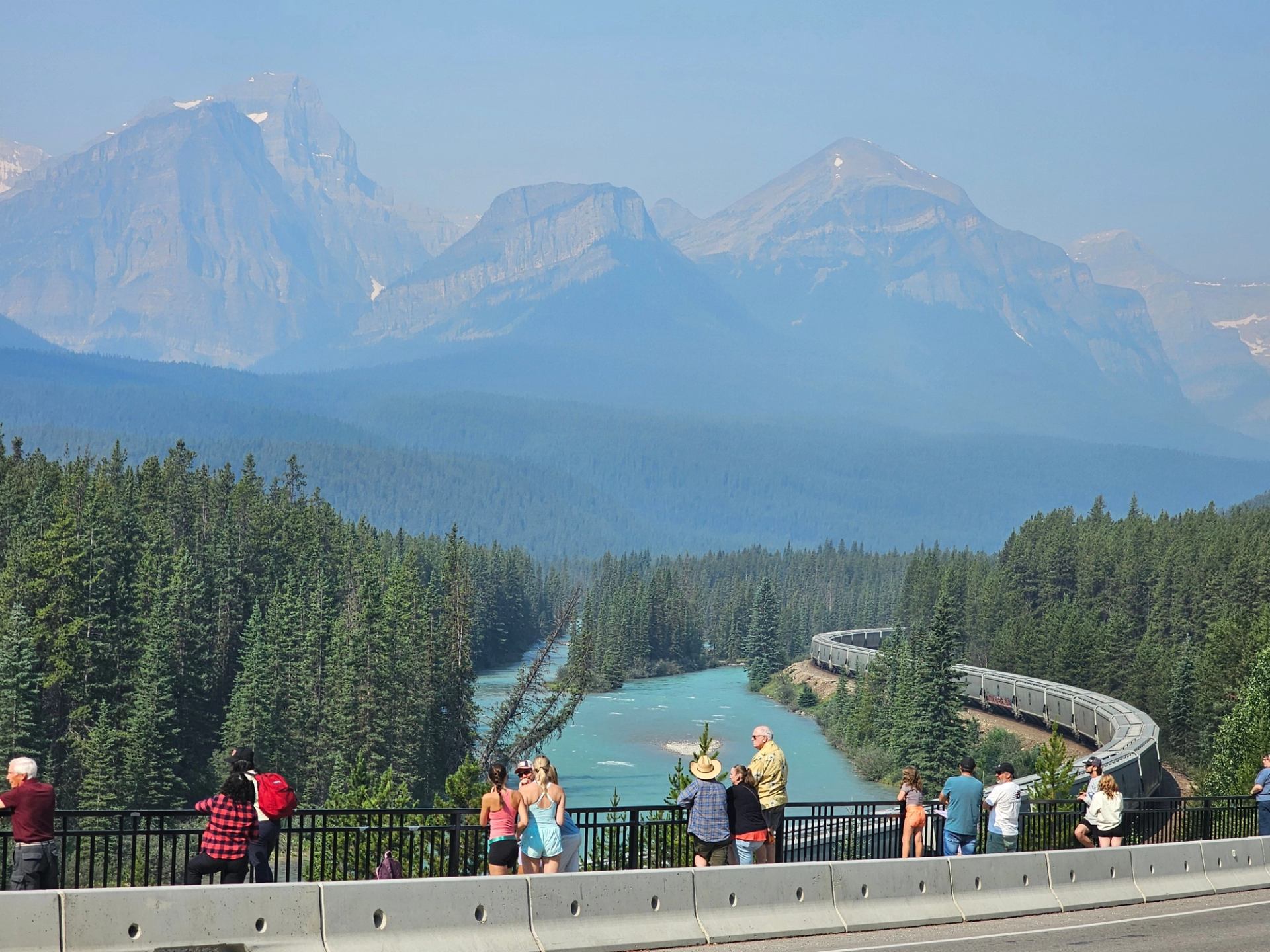 Tourists on roadside viewpoint watch freight train curve beside turquoise river and forested mountains.