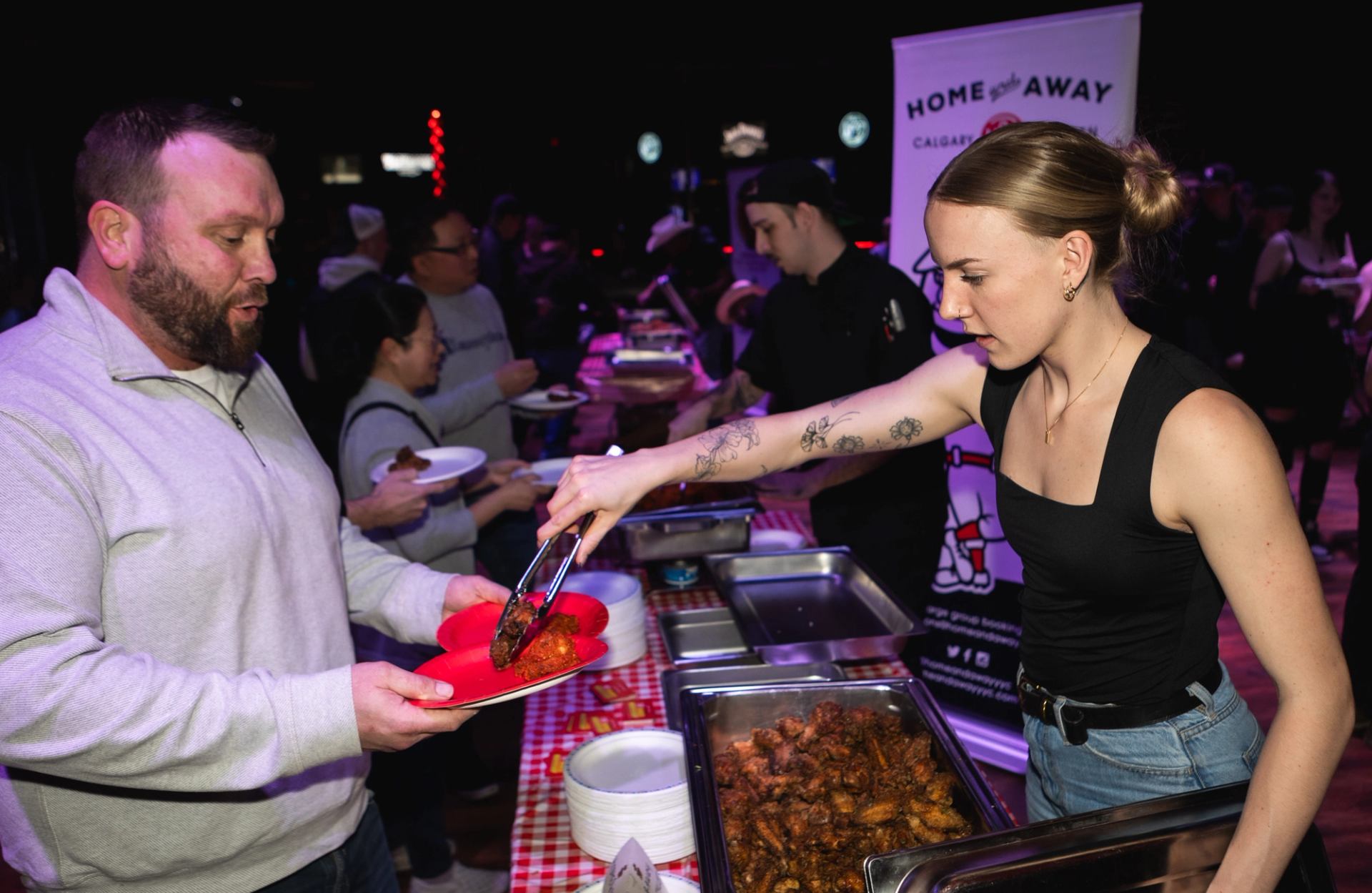 A server places sauced wings onto a guest’s plate at a busy tasting station.