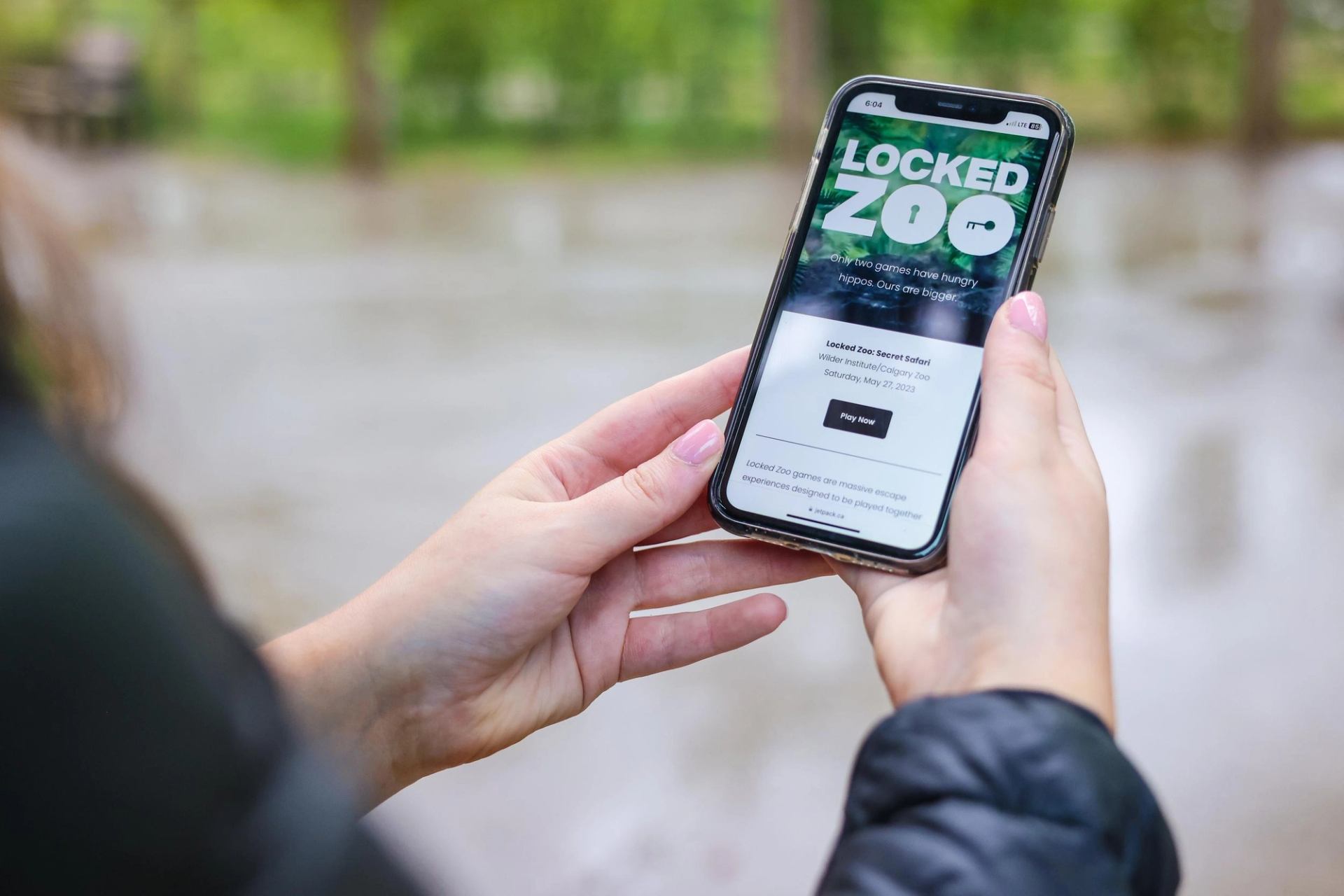 Hands holding smartphone displaying Locked Zoo game webpage outdoors near blurred greenery.