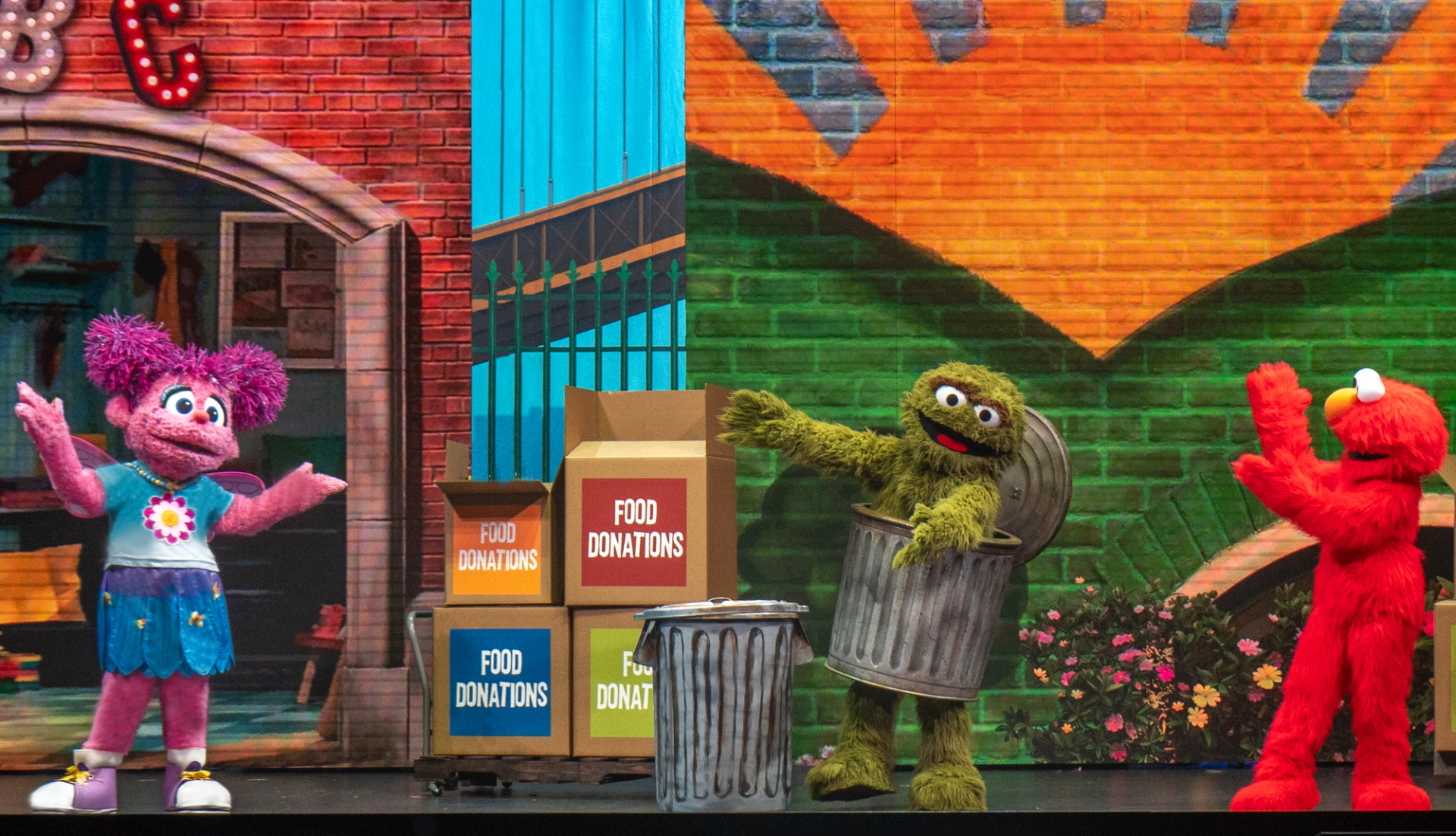 Sesame Street characters stand onstage near food donation boxes and metal garbage cans.