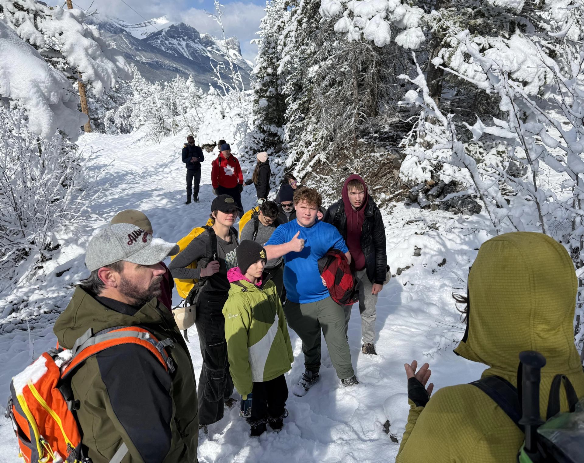 Hikers gather on a snow-covered forest trail beneath rugged mountains.
