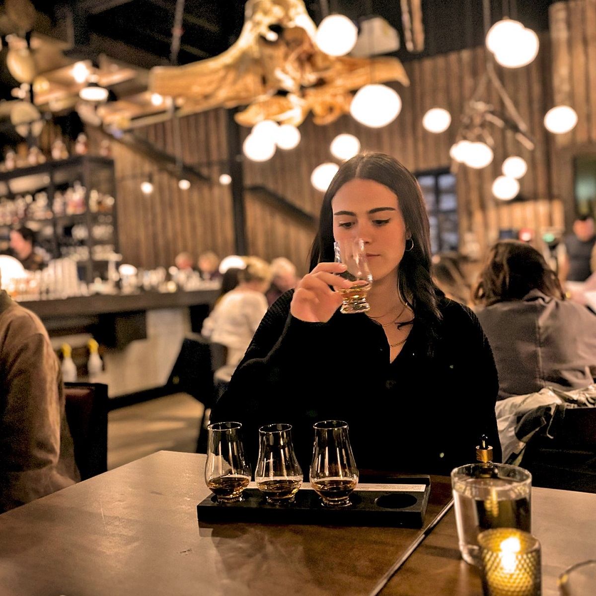 Person tasting a drink in a warmly lit bar with wooden decor and hanging lights.