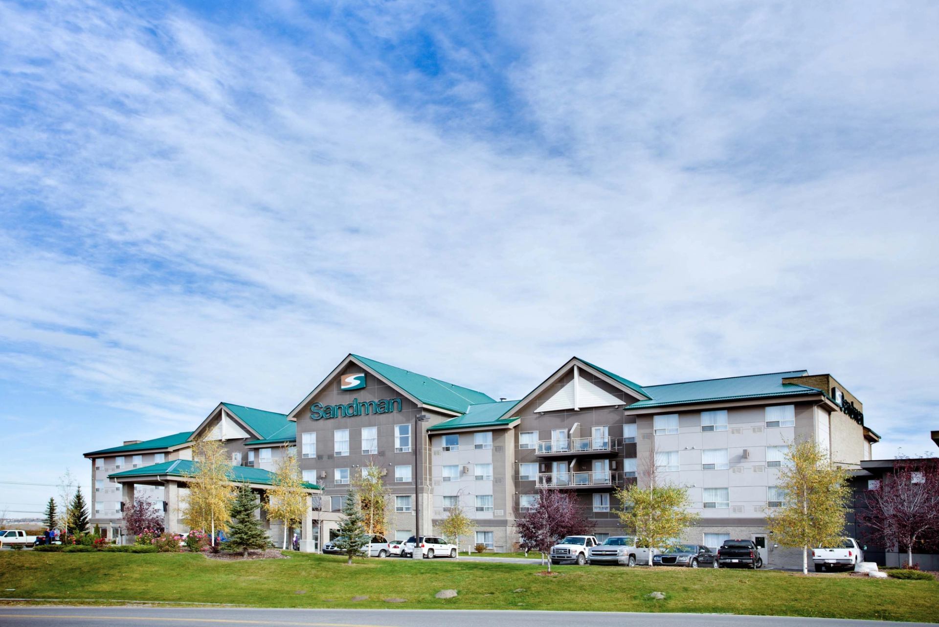 Sandman hotel exterior with teal roofs, parked vehicles and trees under a cloudy sky.