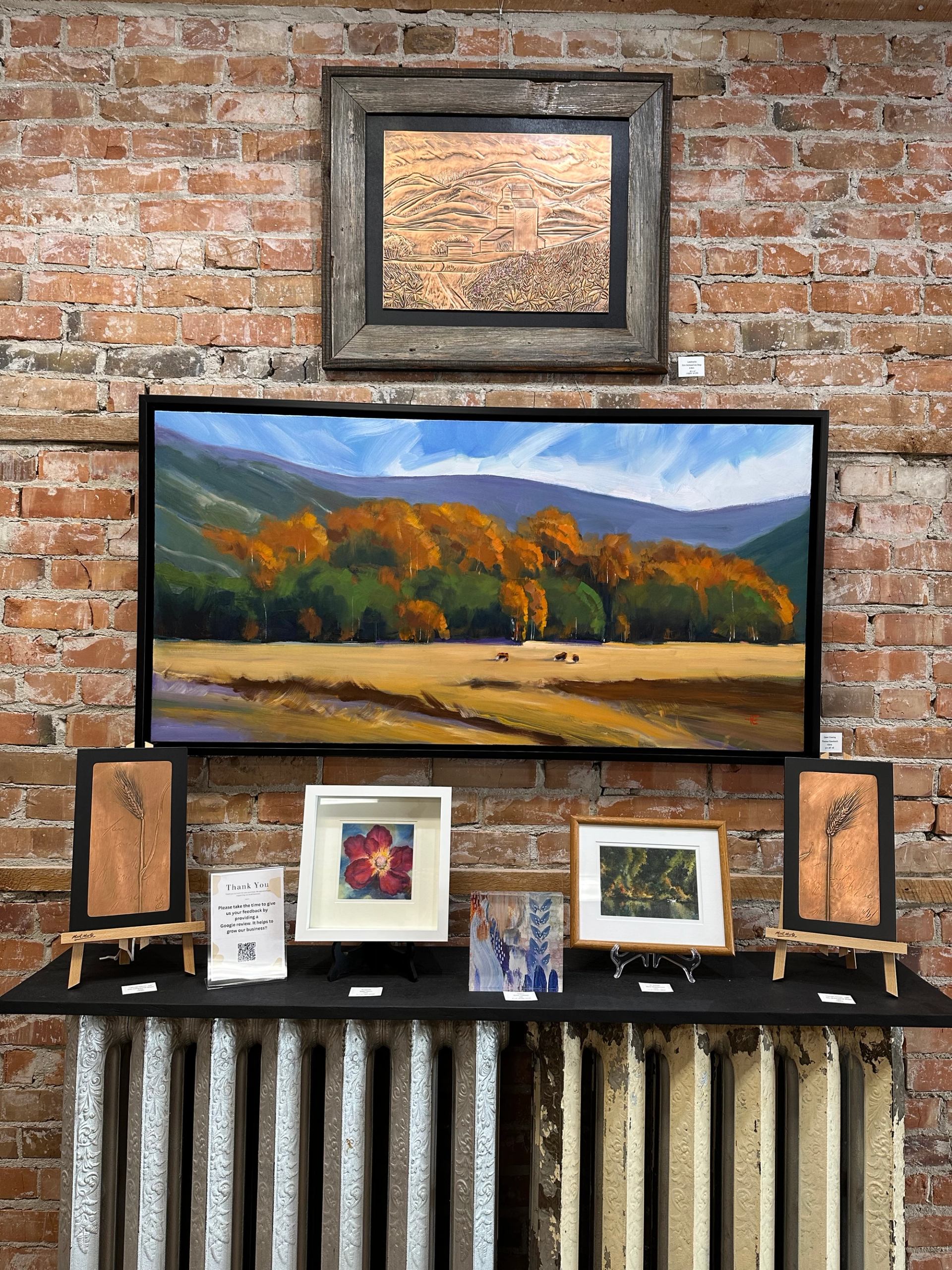 Framed landscape paintings and small artworks arranged on a brick gallery wall.