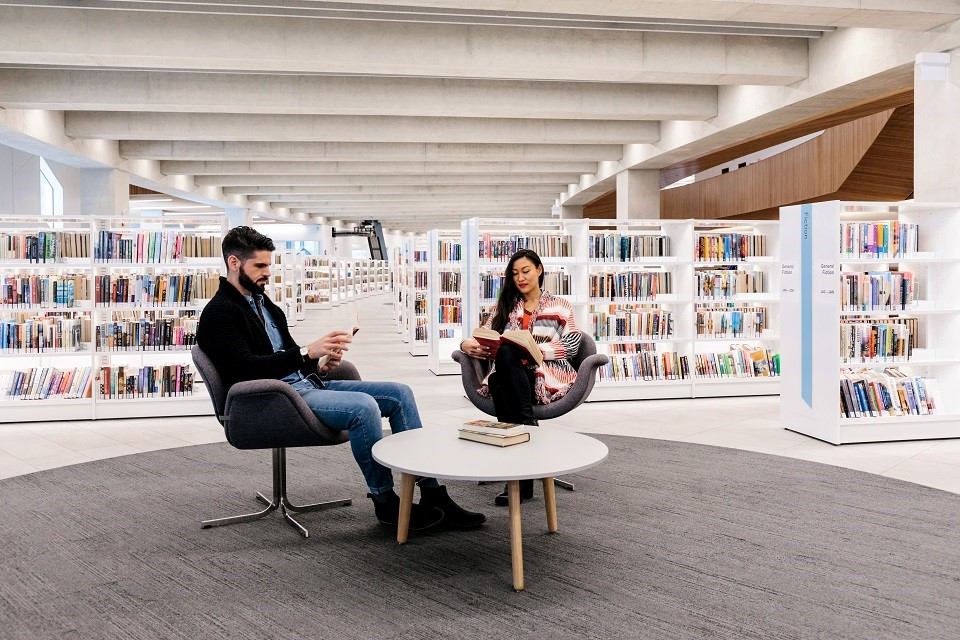 Two people reading in a modern library lounge surrounded by shelves of books.