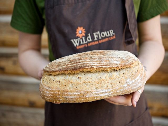 A person wearing a Wild Flour apron holds a round rustic loaf of bread with a golden, crusty exterior. The background shows a warm, wooden setting that highlights a bakery atmosphere.