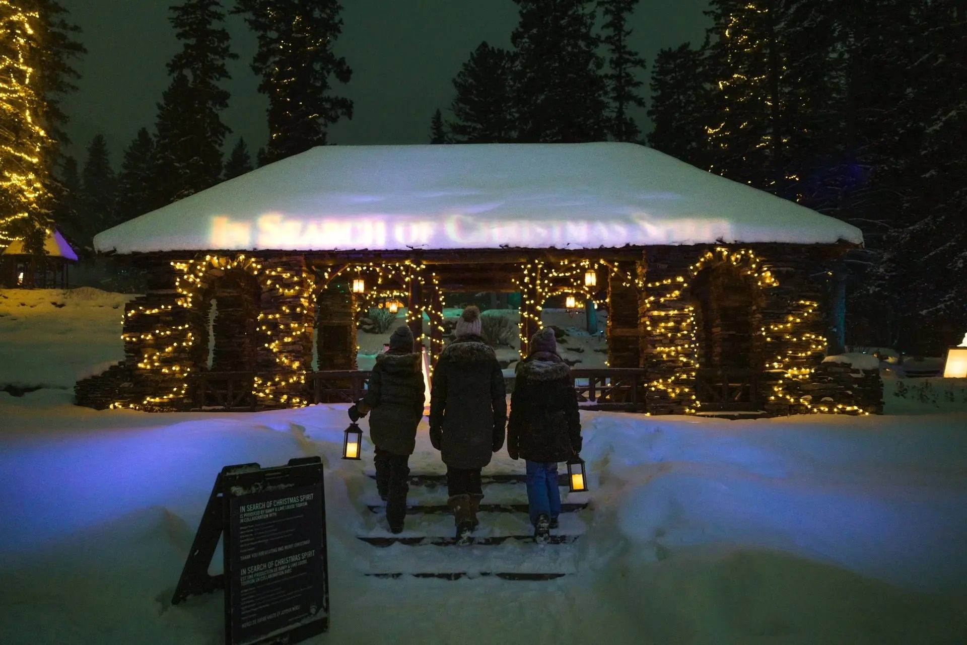 Three people stand by a lit cabin in snowy woods, surrounded by festive Christmas lights.