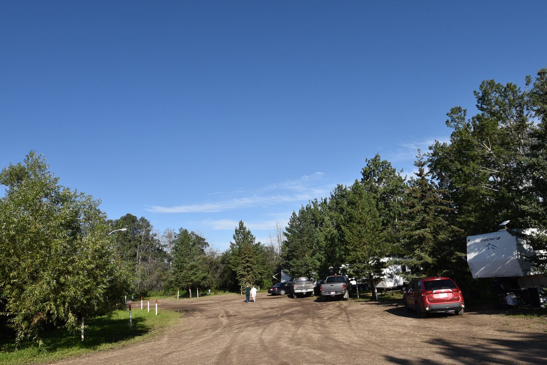 Gravel road in RV park with parked trucks, trailers and people walking among trees.
