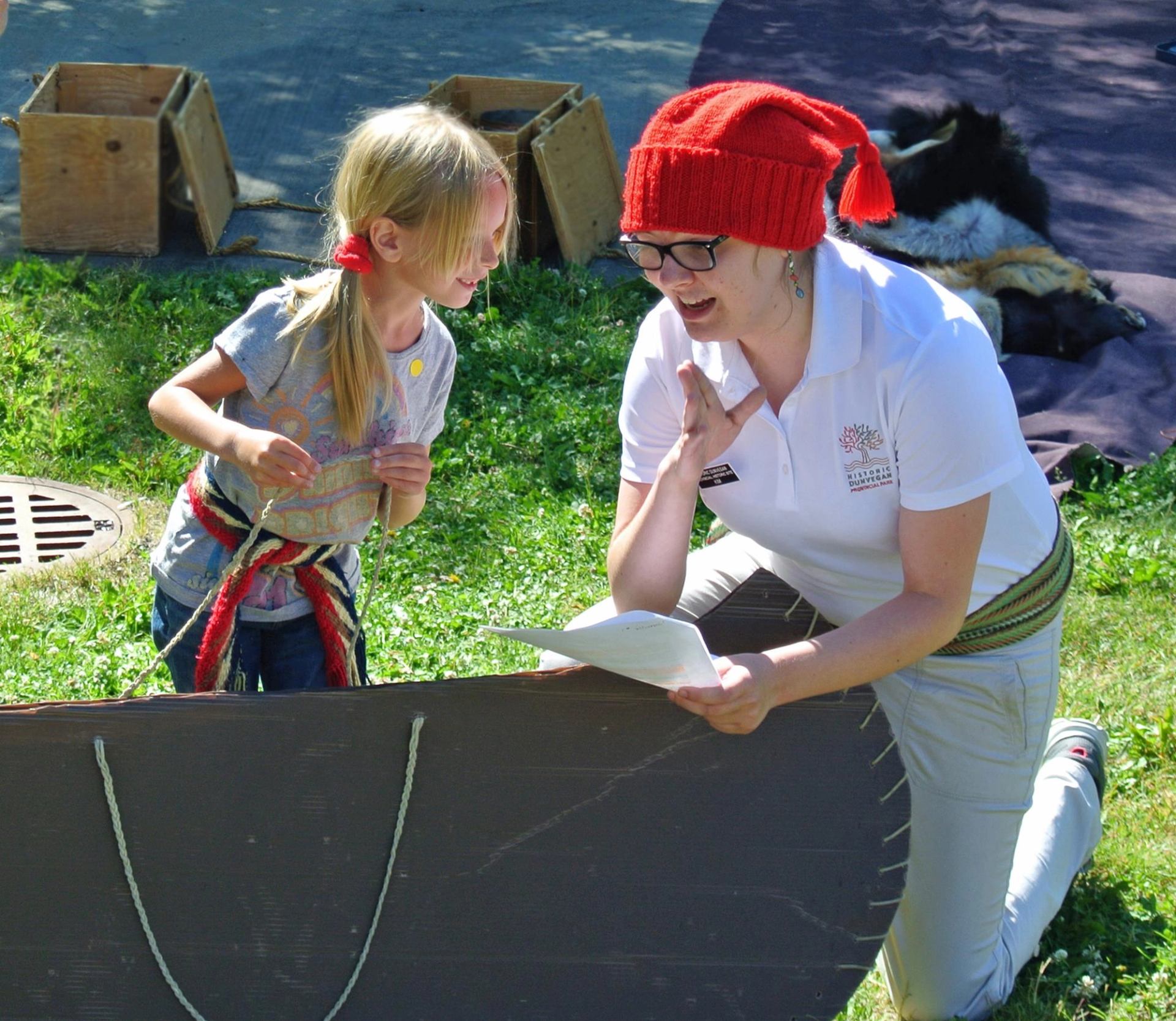 A woman in a red hat and a young girl, both wearing sashes, interact enthusiastically around a boat-like structure outdoors.