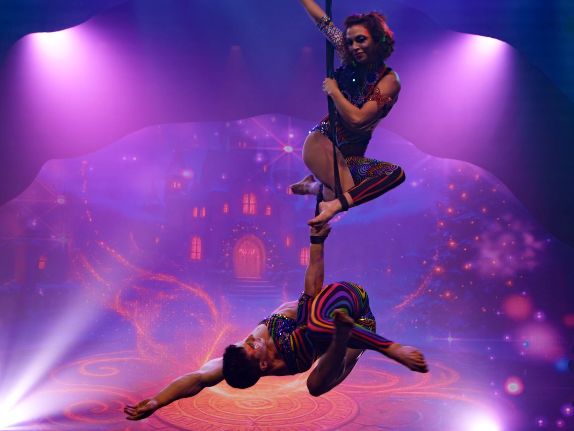 Aerial acrobats balance vertically on a suspended strap under purple stage lights.