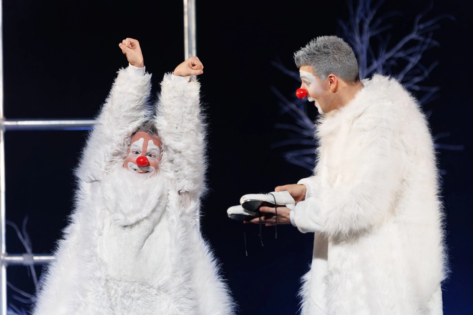 Two clowns in white furry costumes perform onstage, one raising arms, one holding shoes.