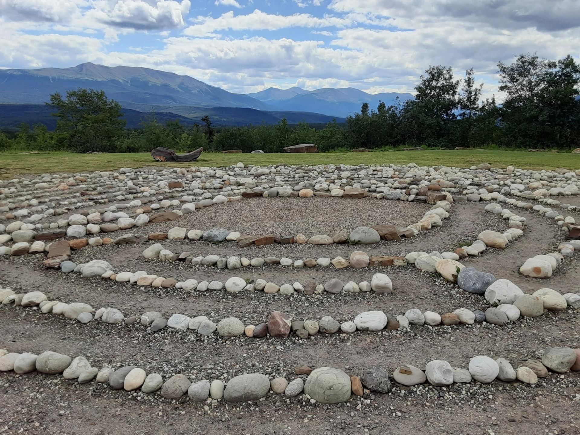 Stone labyrinth on gravel with distant tree line and mountain range under clouds.