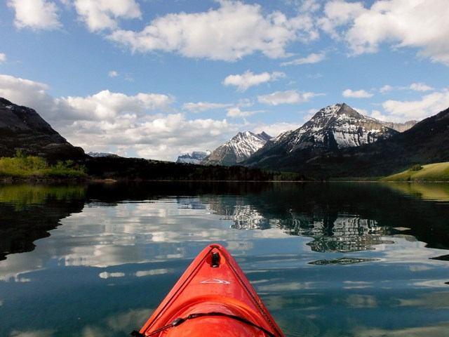 Front of red kayak on calm lake with mountains in the background.