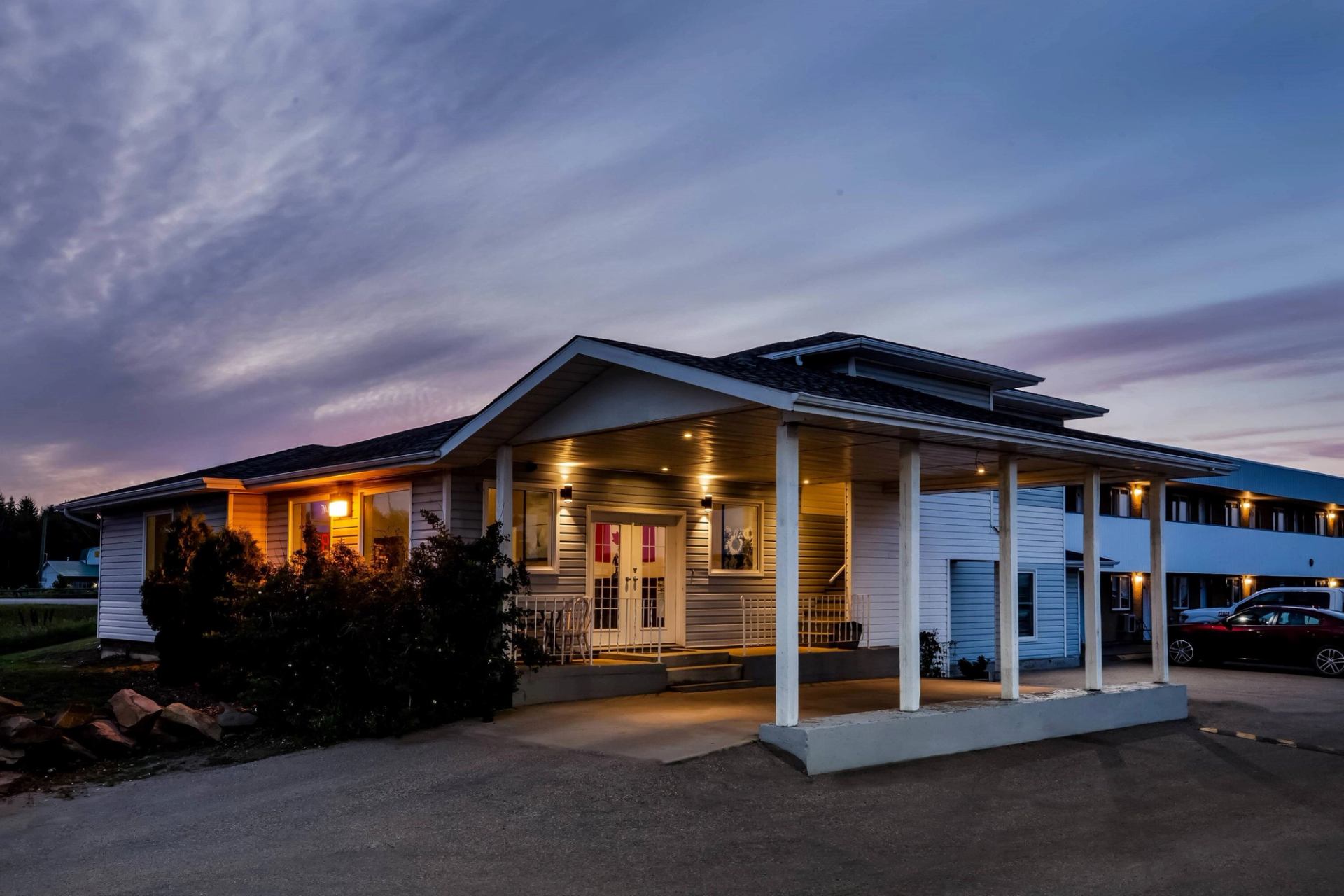 The hotel with lights glowing around the entrance and rooms, set against a twilight sky. A car is parked nearby, and the driveway is clear.
