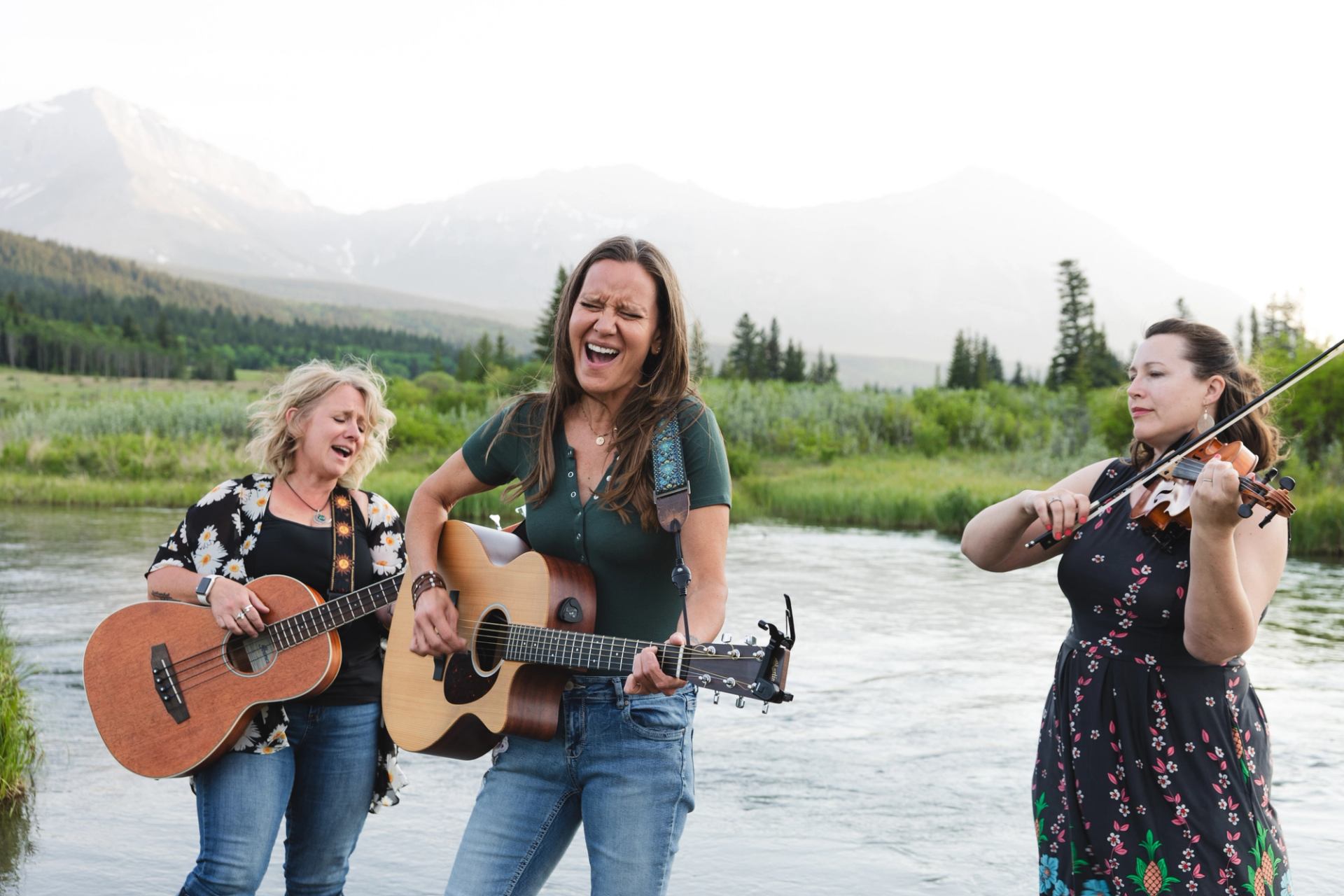 Three women playing acoustic guitars and a violin by a river with mountains.