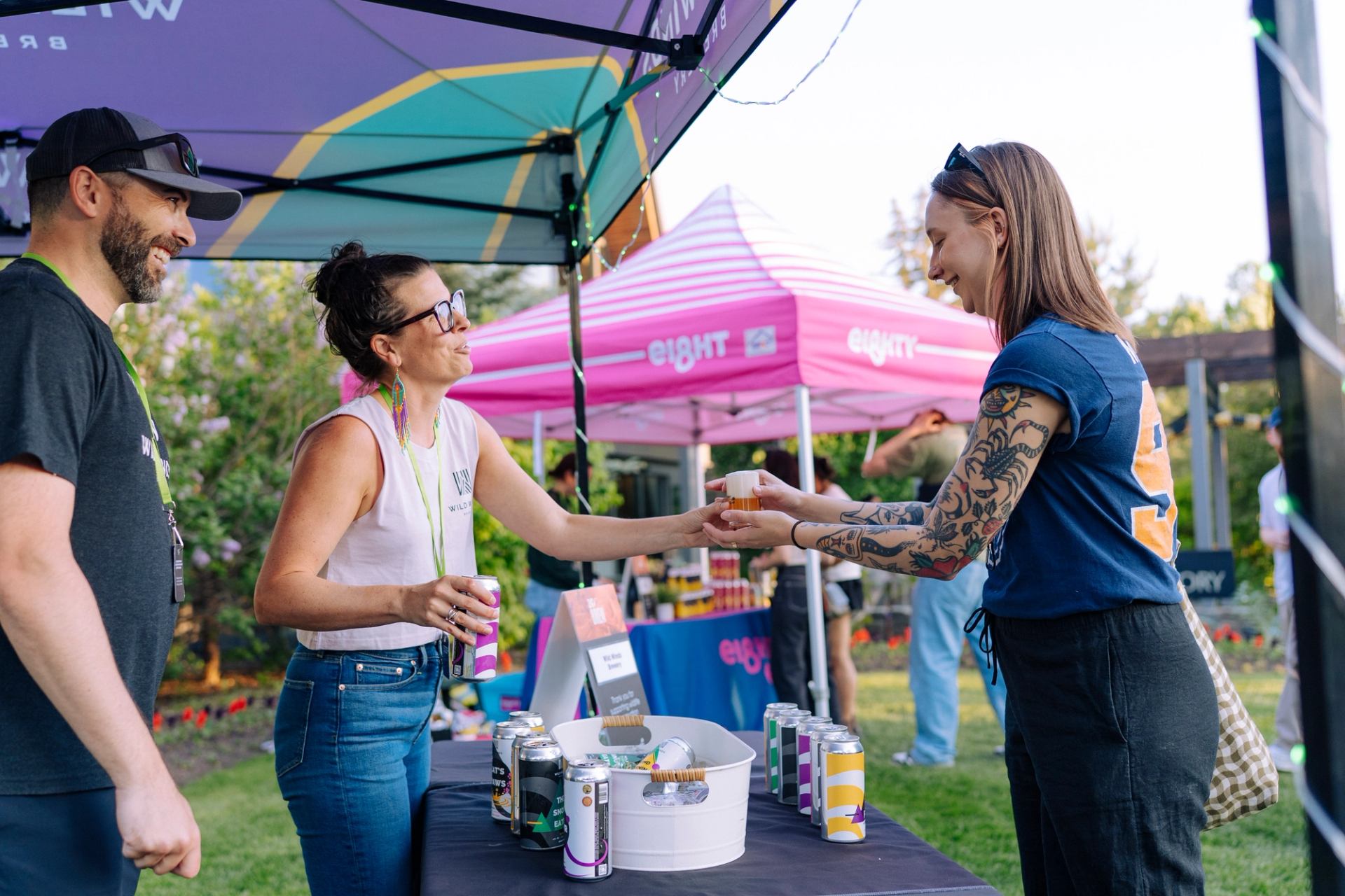 Attendees sample craft drinks at a vendor booth under tents at an outdoor event.