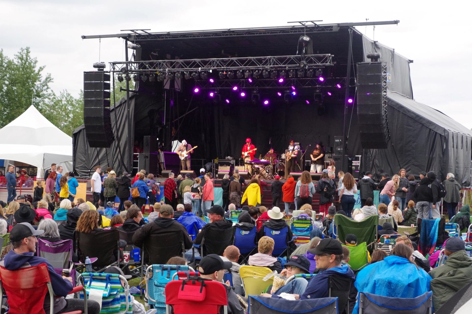 Music stage with purple lights, band performing, and audience seated in festival chairs.