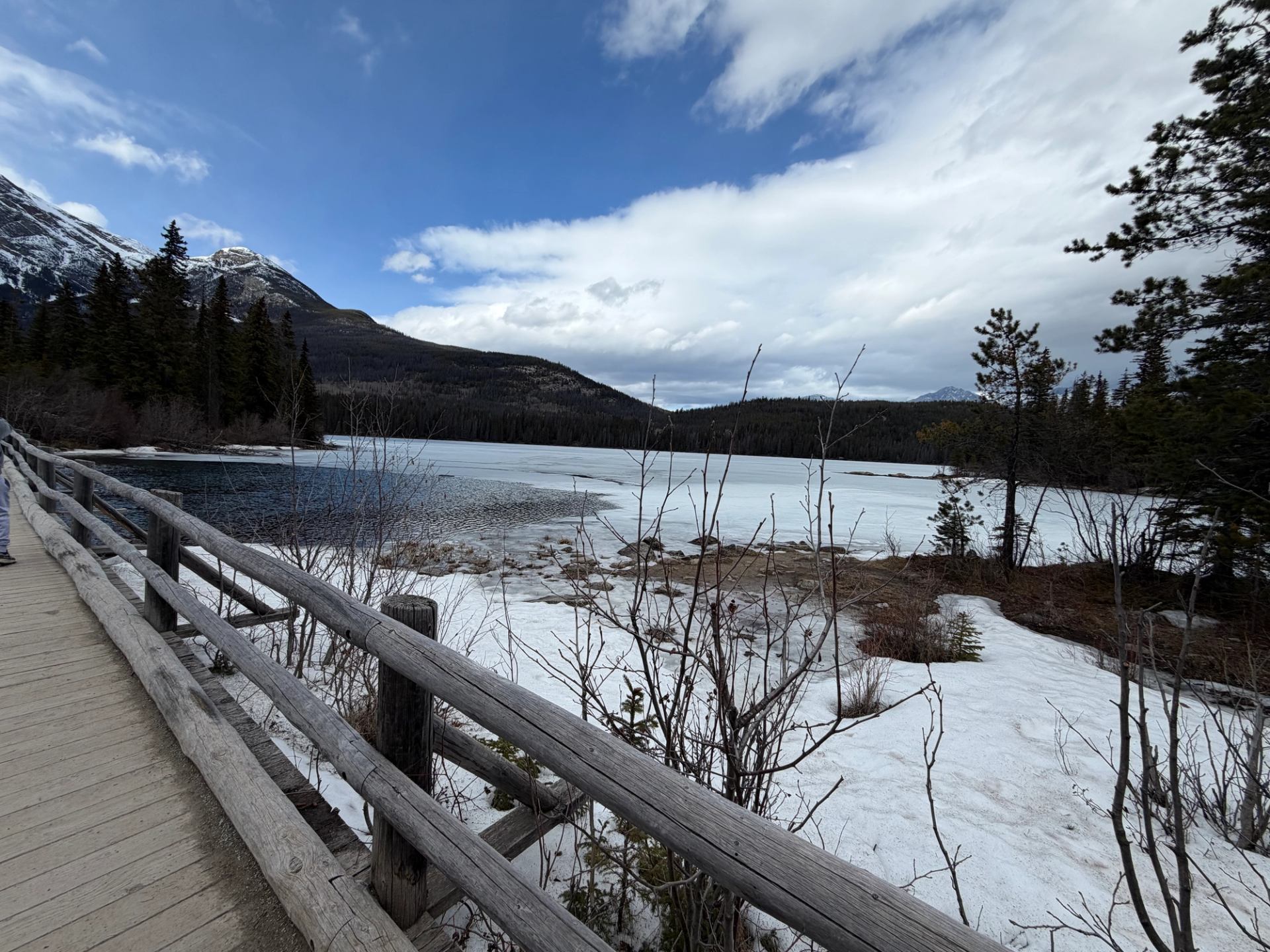 Wooden boardwalk beside partially frozen lake, with snow, evergreen trees and distant mountains.
