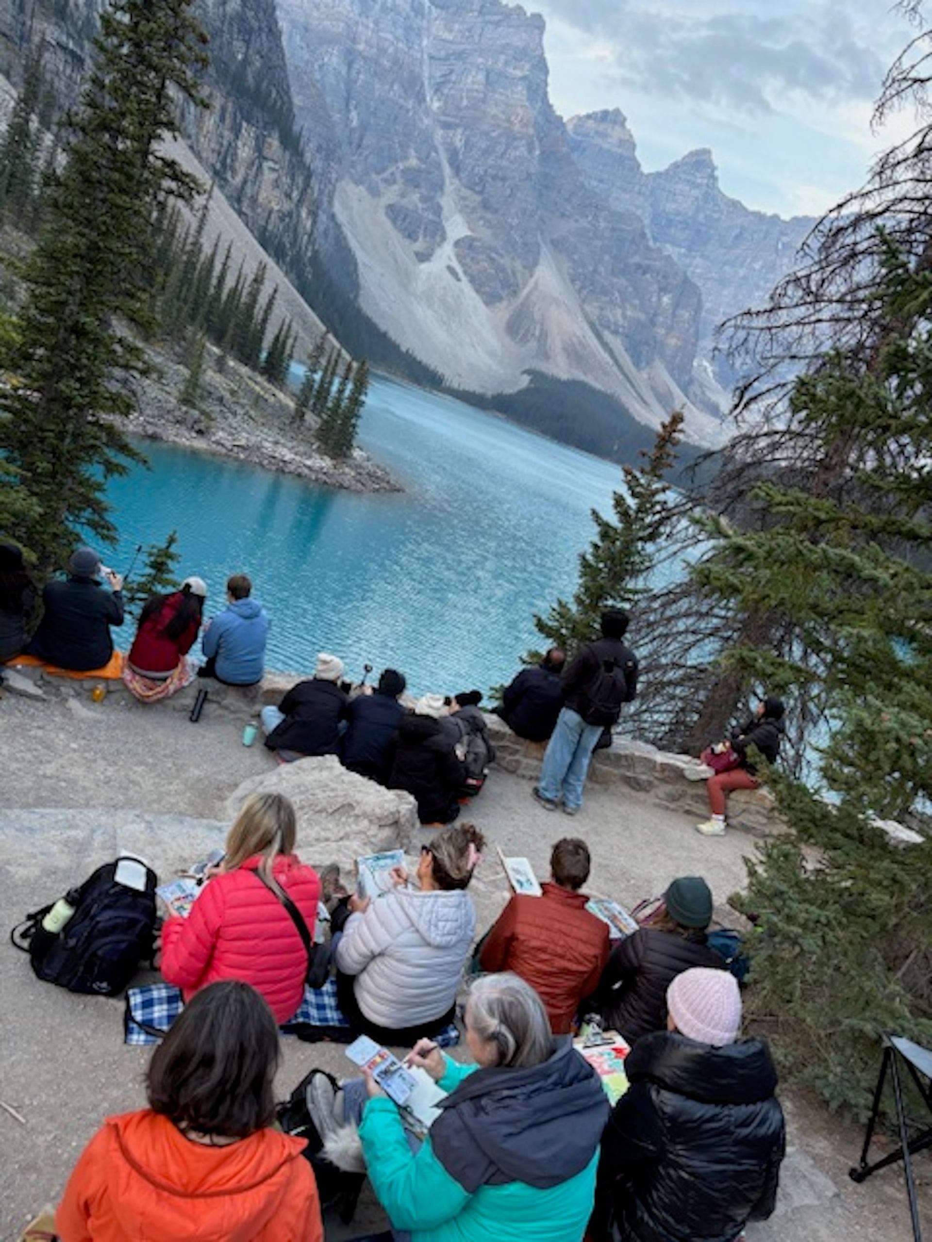 Group of people sketching beside turquoise mountain lake surrounded by steep rocky peaks.