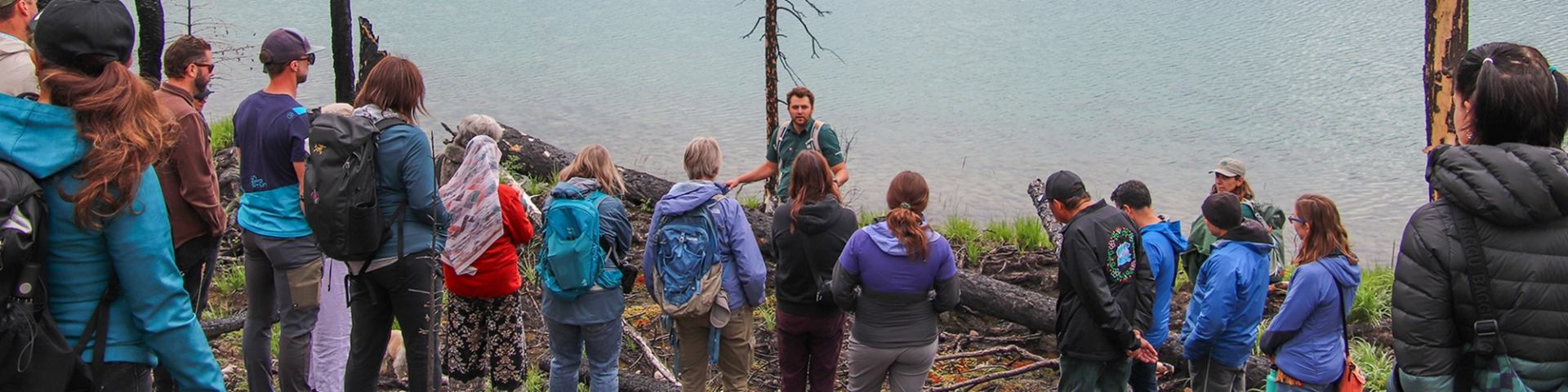 Outdoor group listens to guide speaking beside lakeshore and fallen trees on grassy slope.