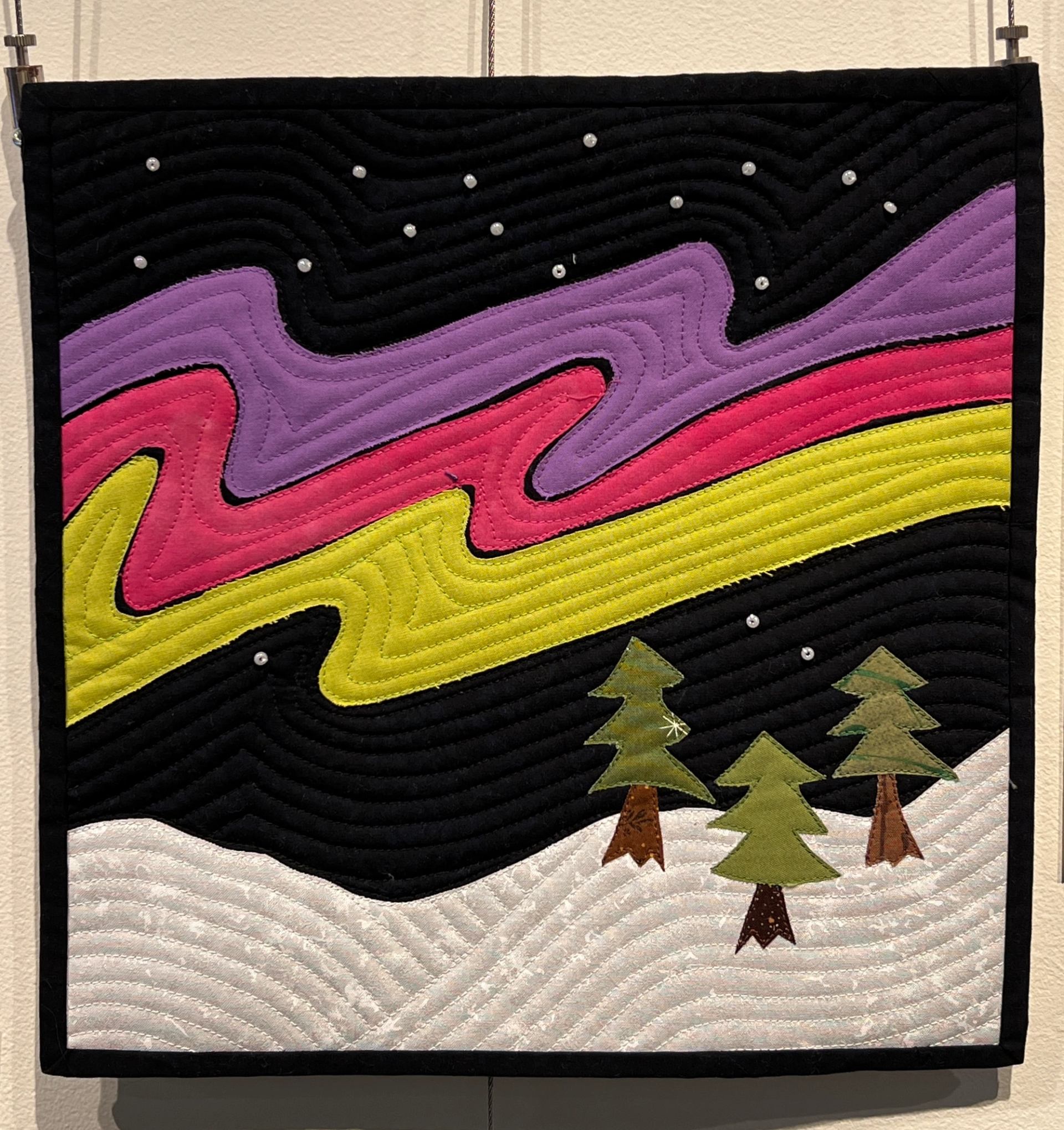 A quilted scene featuring purple, pink, and yellow Northern Lights against a starry black sky, above snow-covered ground with three green evergreen trees.