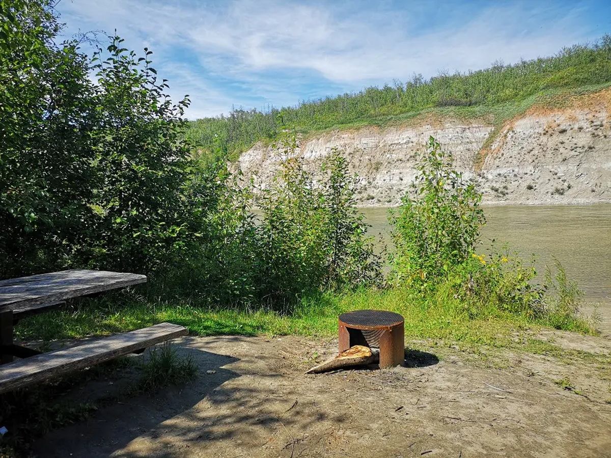 Riverside campsite with picnic table, metal fire pit, shrubs and eroded cliffside.