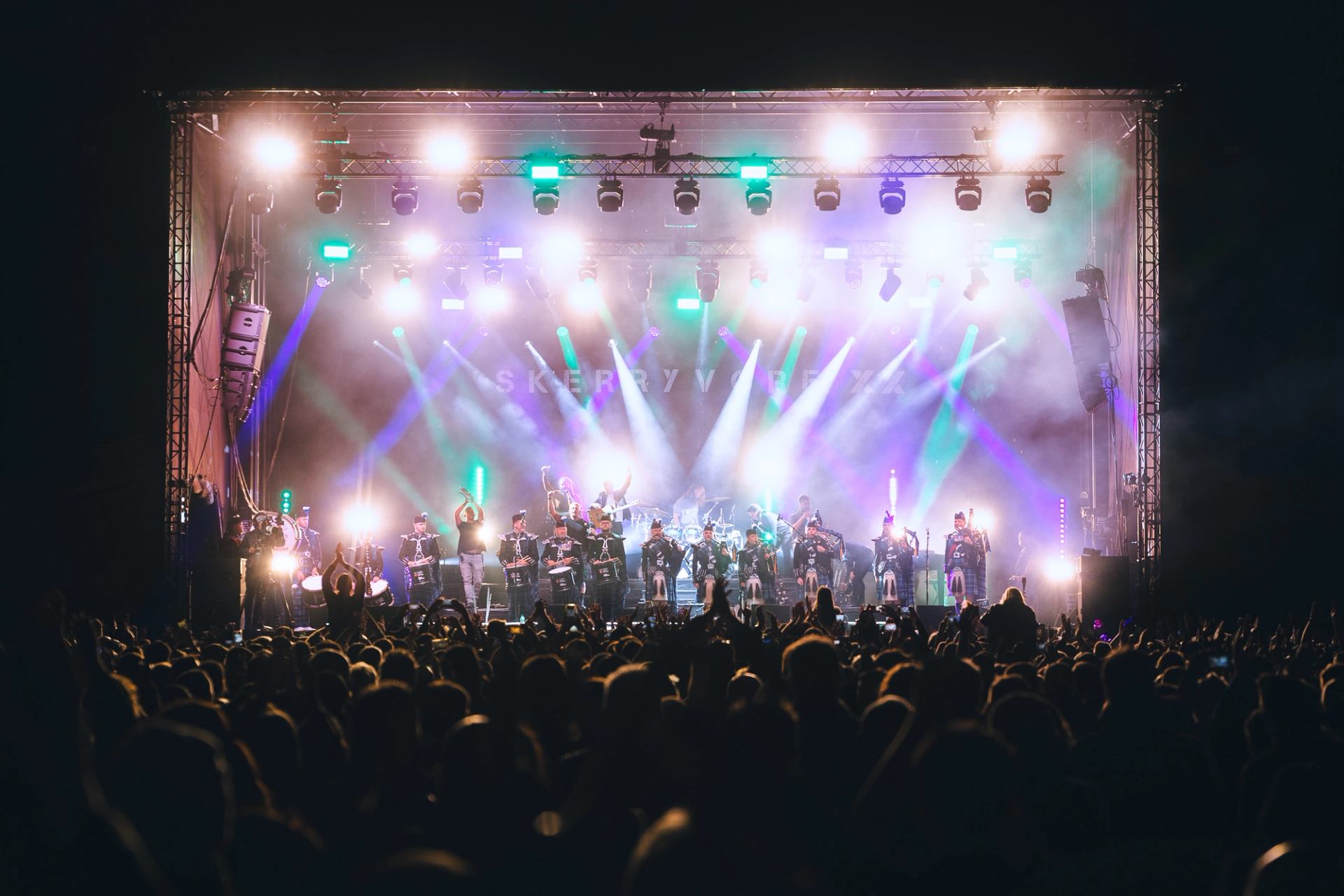 Large crowd watches musicians perform on brightly lit outdoor stage with colourful beams.